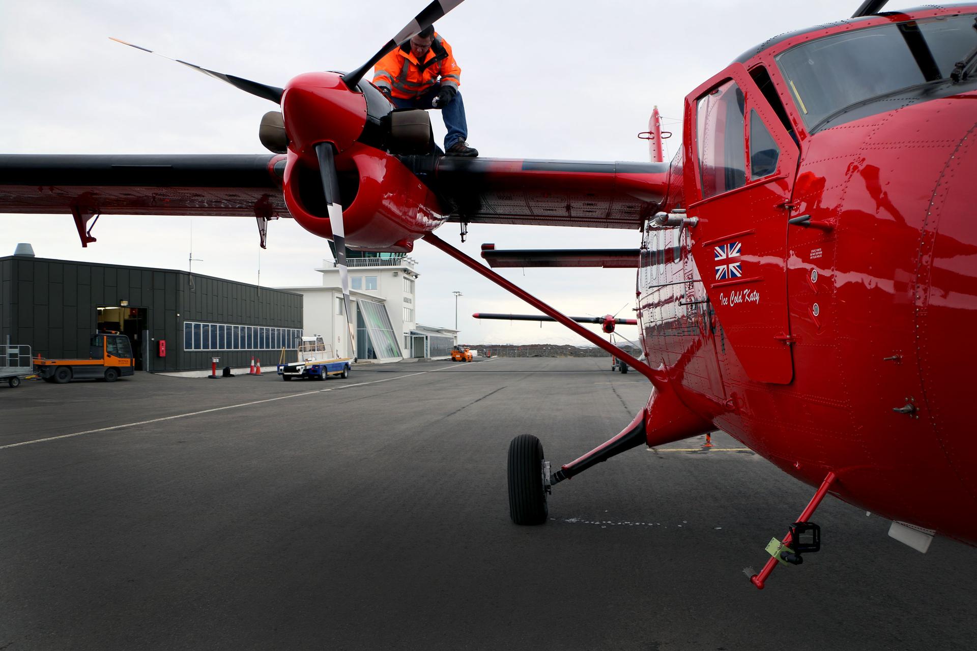 A man is perched atop a propeller of a small twin engine plane on a tarmac. The plane is bright cherry red.