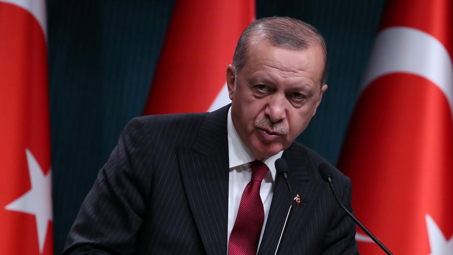 Turkish President Tayyip Erdoğan is shown speaking at a podium with Turkish flags behind him.