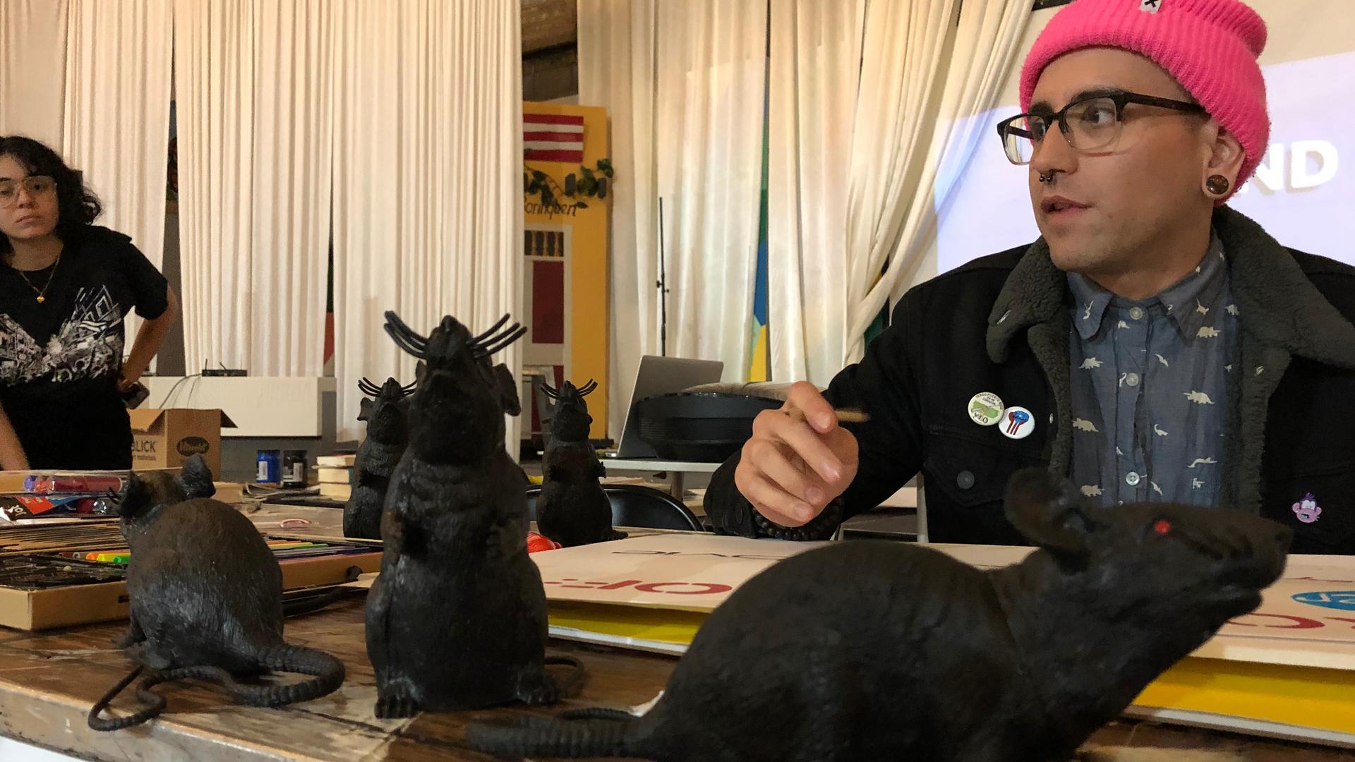 A person wearing a pink cap sits at a table near three large, black, plastic rats.