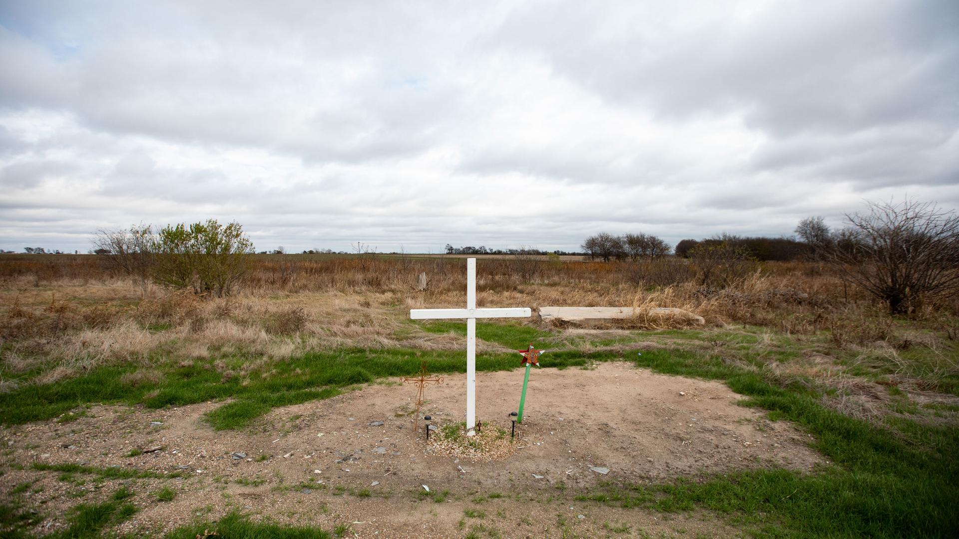A single white cross is shown on a small dirt area amongst a wide open grassy field.