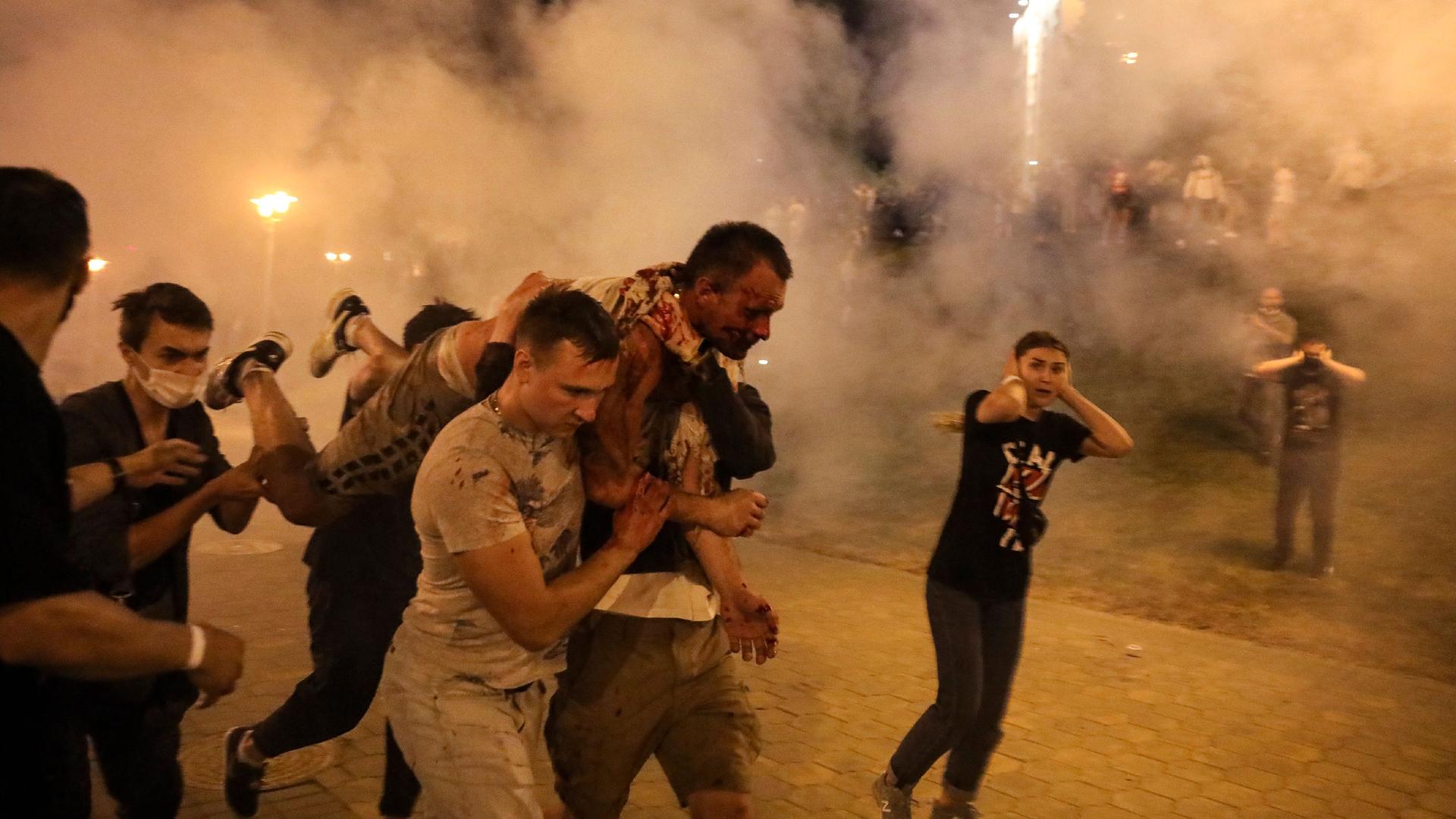 Several people are shown carrying an injured protester on their shoulders with smoke all around them.