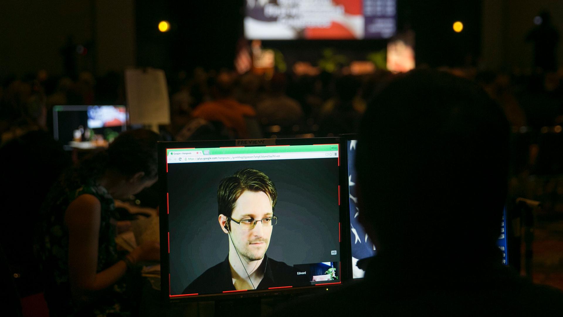 A small computer screen is shown in a dark room with Edward Snowden appearing in a close-up frame.