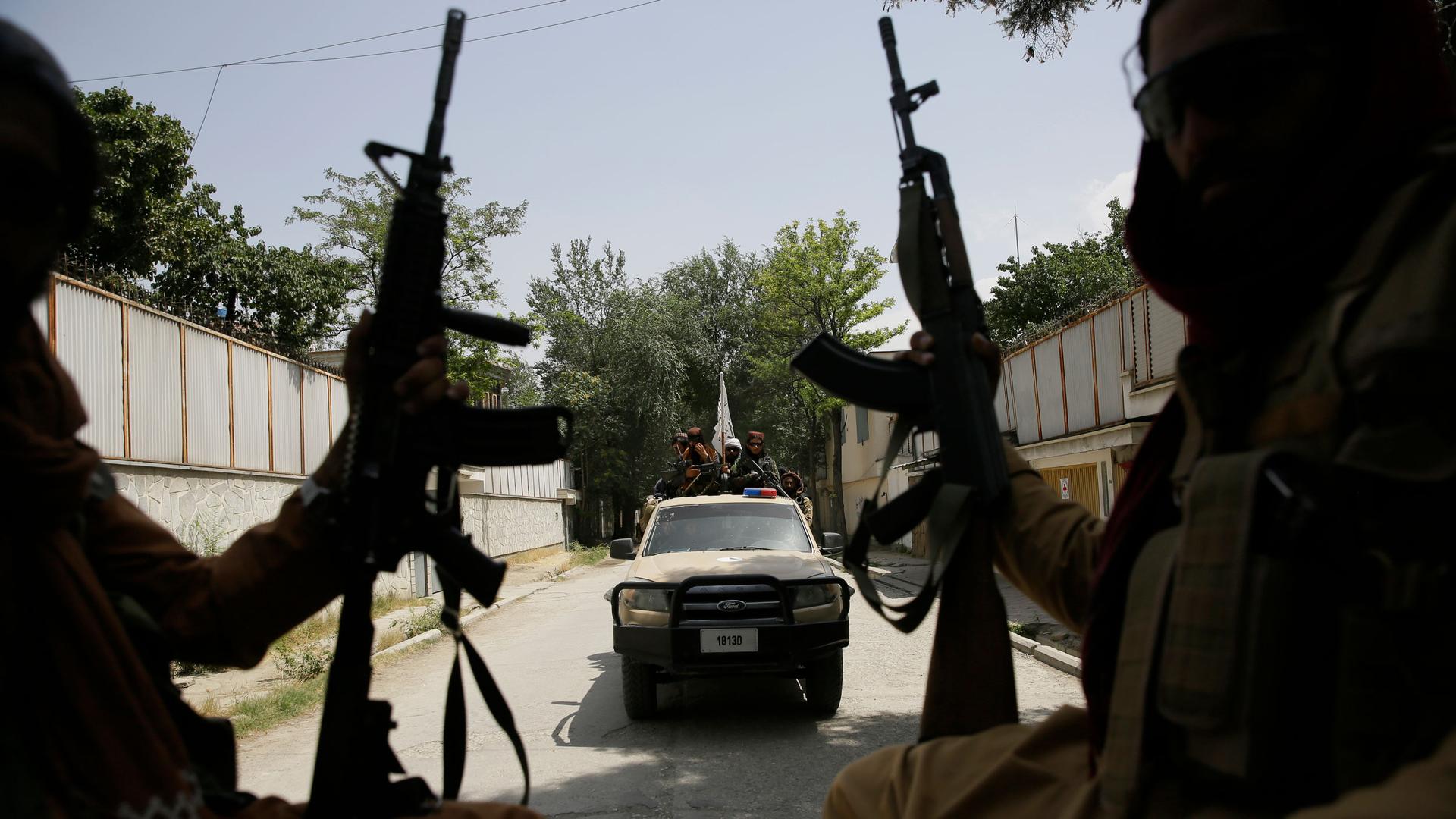 A pickup truck is shown flying the white and black Taliban flag in the distance with two people holding rifles in the nearground.