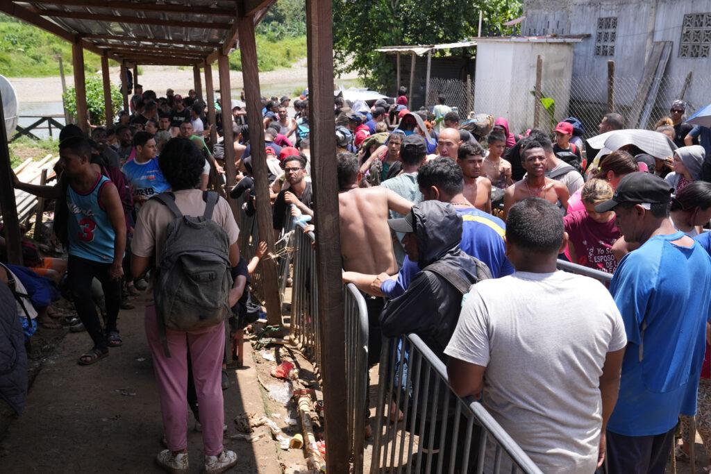 A crowd of people push up against a metal gate while waiting in a line wearing backpacks and street clothes.