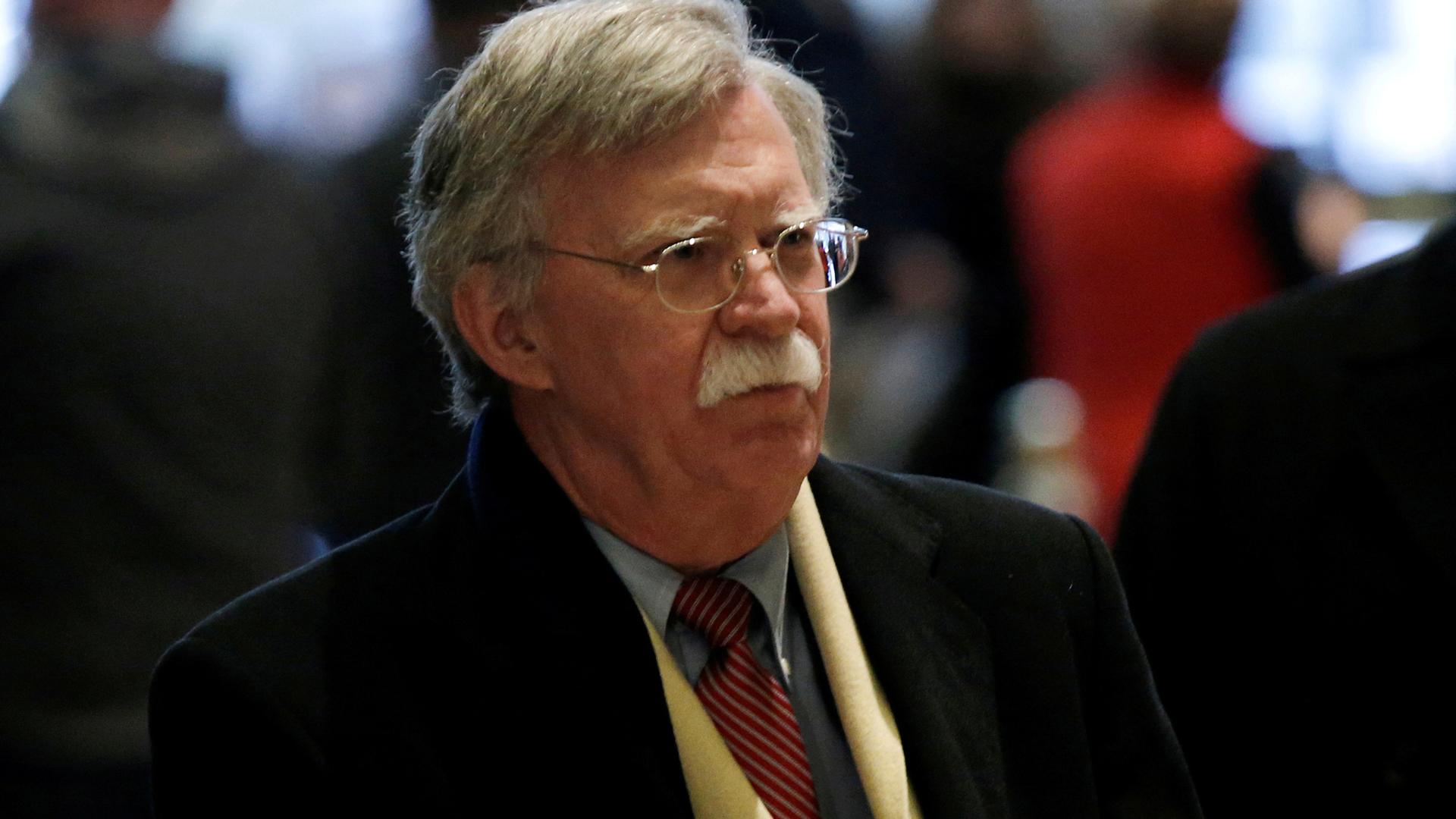 Former US Ambassador to the United Nations John Bolton walks through a lobby wearing a scarf and overcoat.