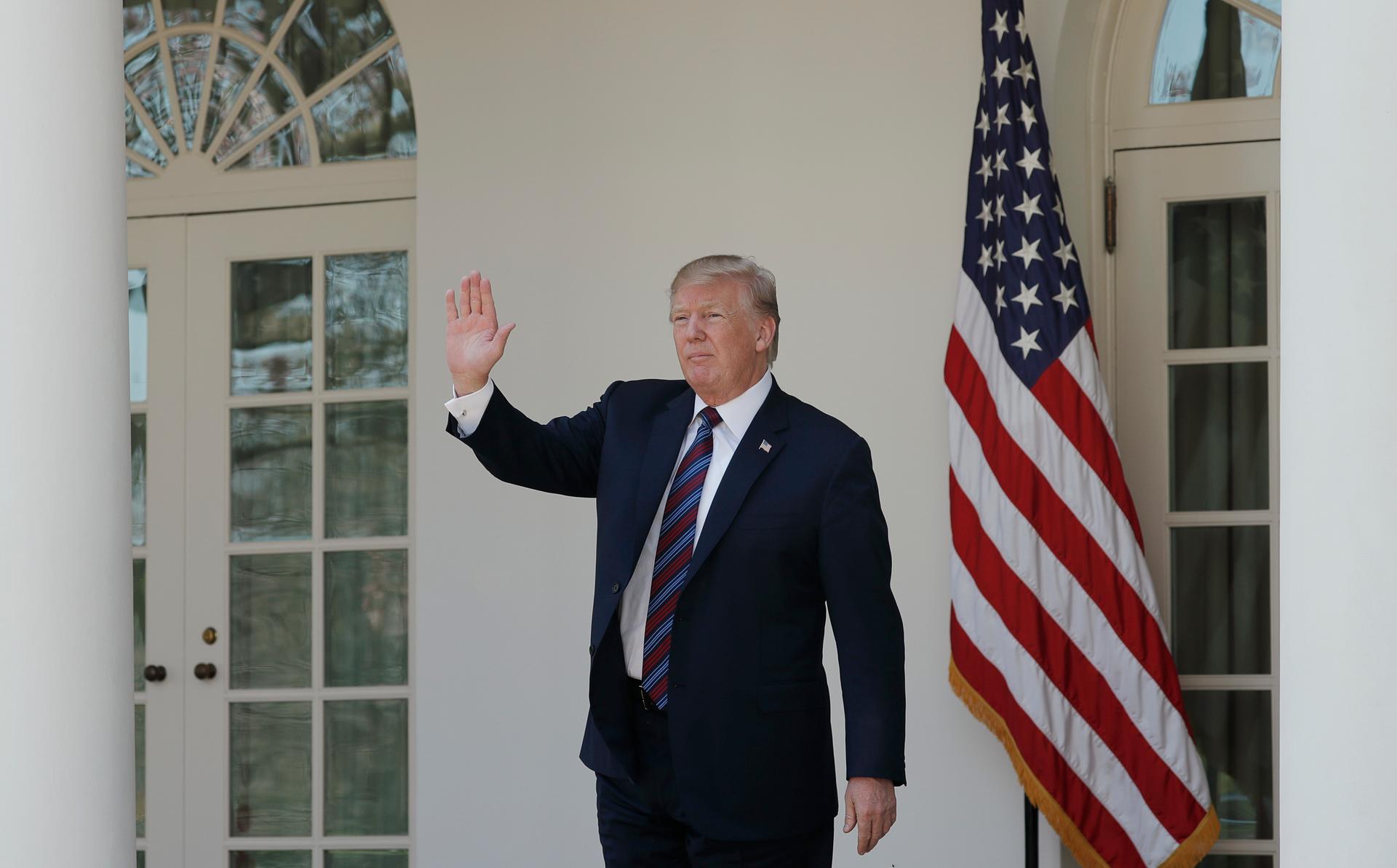 President Donald Trump, in a suit with the White House  behind him waves in the Rose Garden.