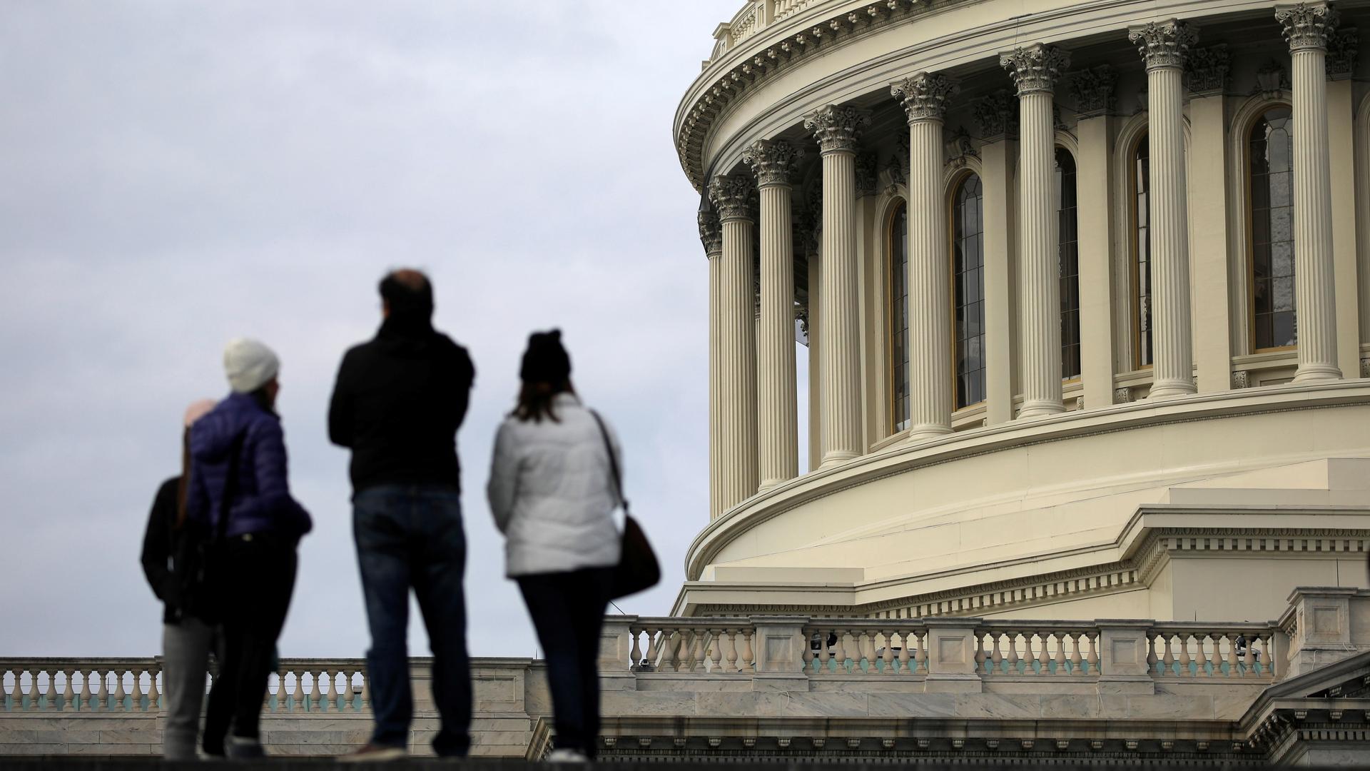 Tourists stand in the foreground out of focus with the US Capitol building in focus in the background.