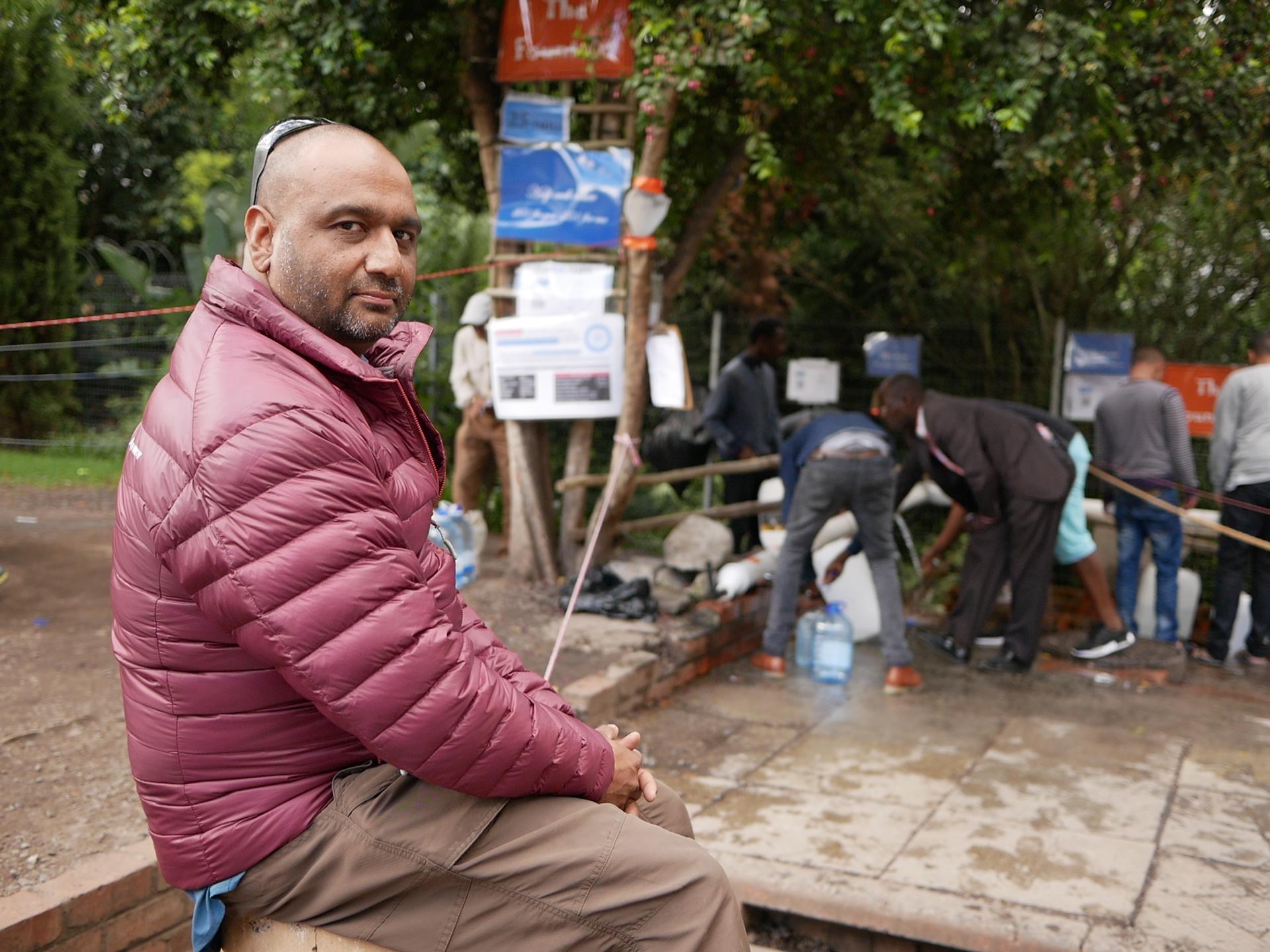 Portrait of Riyaz Rawoot, who wears a red coat as he watches people take their turns filling bottles at a spring in Newlands, Cape Town.