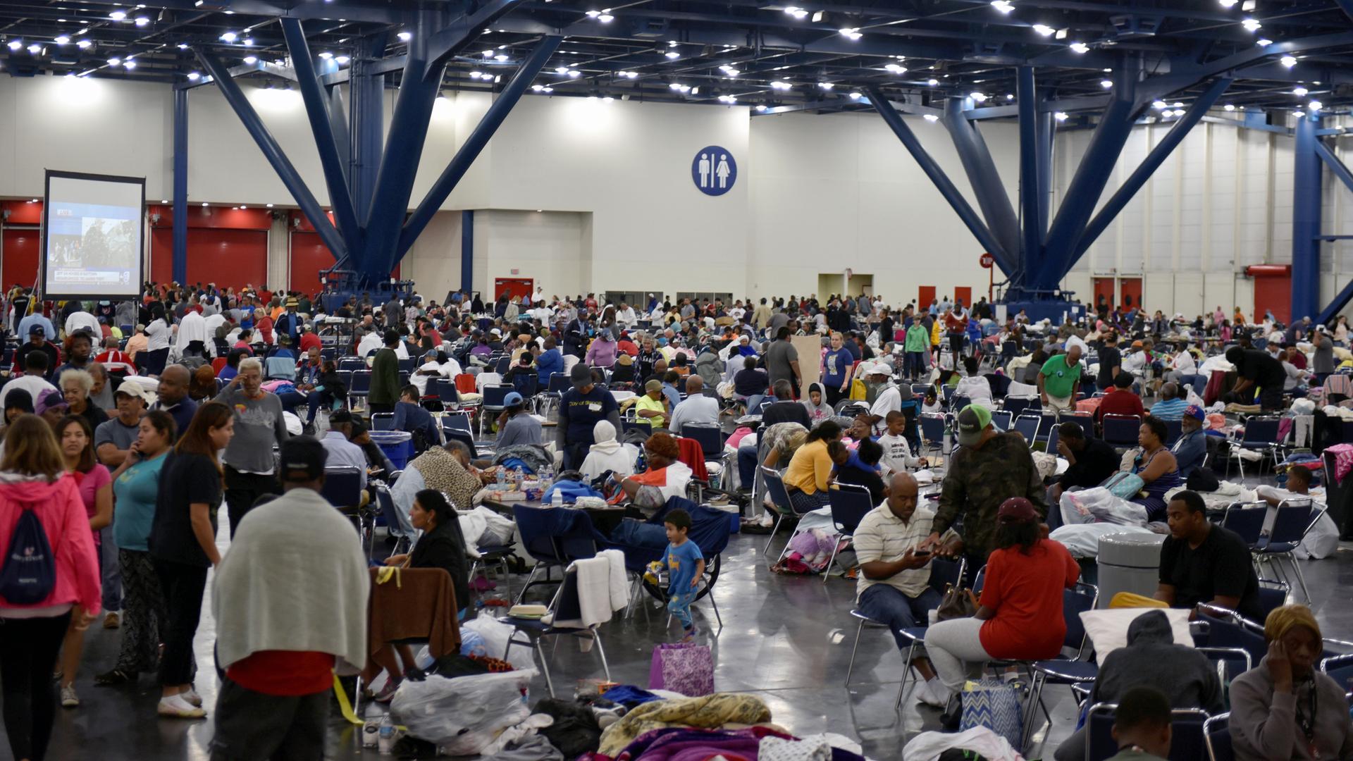 Hundreds of people are gathered in a room. Some are seated at tables and have water bottles, some pillow or blankets with them. A large screen is set up to broadcast news about Tropical Storm Harvey.