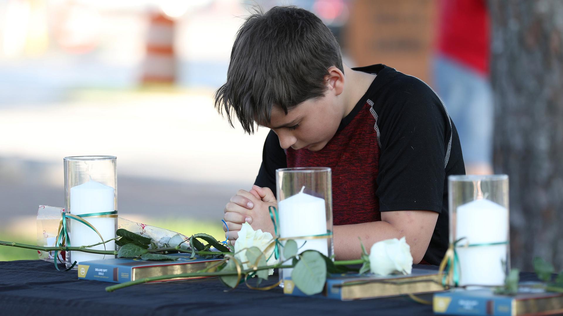 A young boy bends his head in prayer. He is surrounded by several white candles on either side.