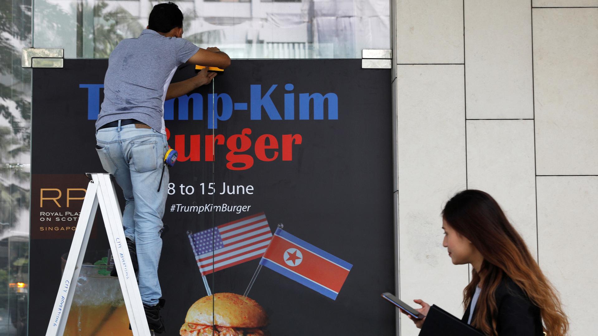 A worker in blue jeans, stands on a ladder and puts up an advertisement for the Trump-Kim burger.