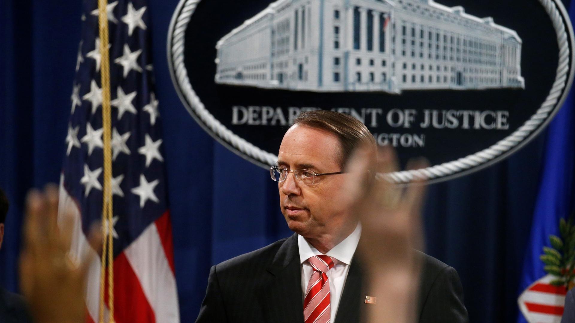 Deputy US Attorney General Rod Rosenstein is seen in front of an emblem of the US Justice Department with reporter's hands raised in the near ground.