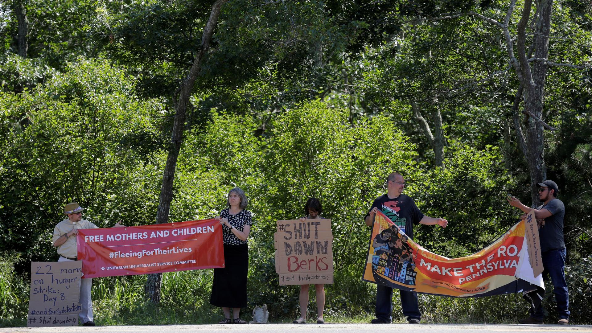 People stand on side of road in front of trees holding signs; one reads "Shut Down Berks"