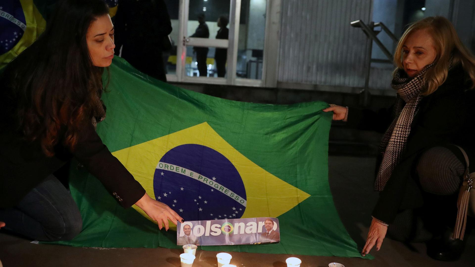 Two women kneel next to a Brazilian flag as they light candles