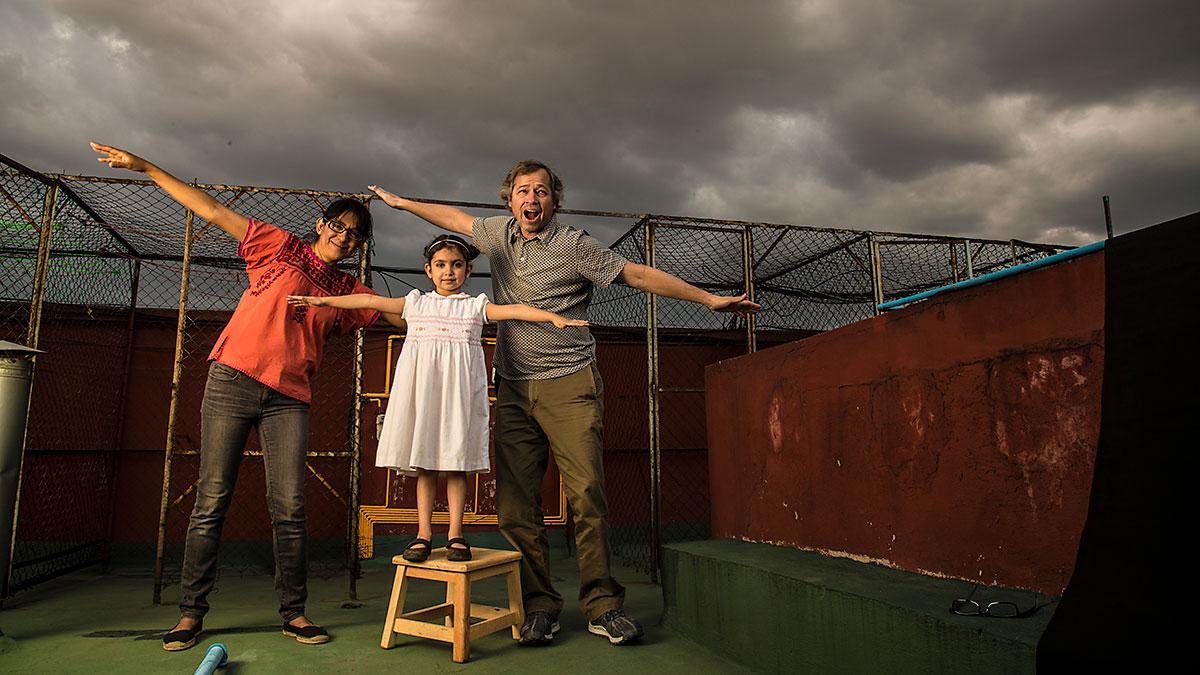 A woman, a child and man spread their arms out like airplanes on the roof of a building as they pose for a silly family photograph. Behind them are dark storm clouds.