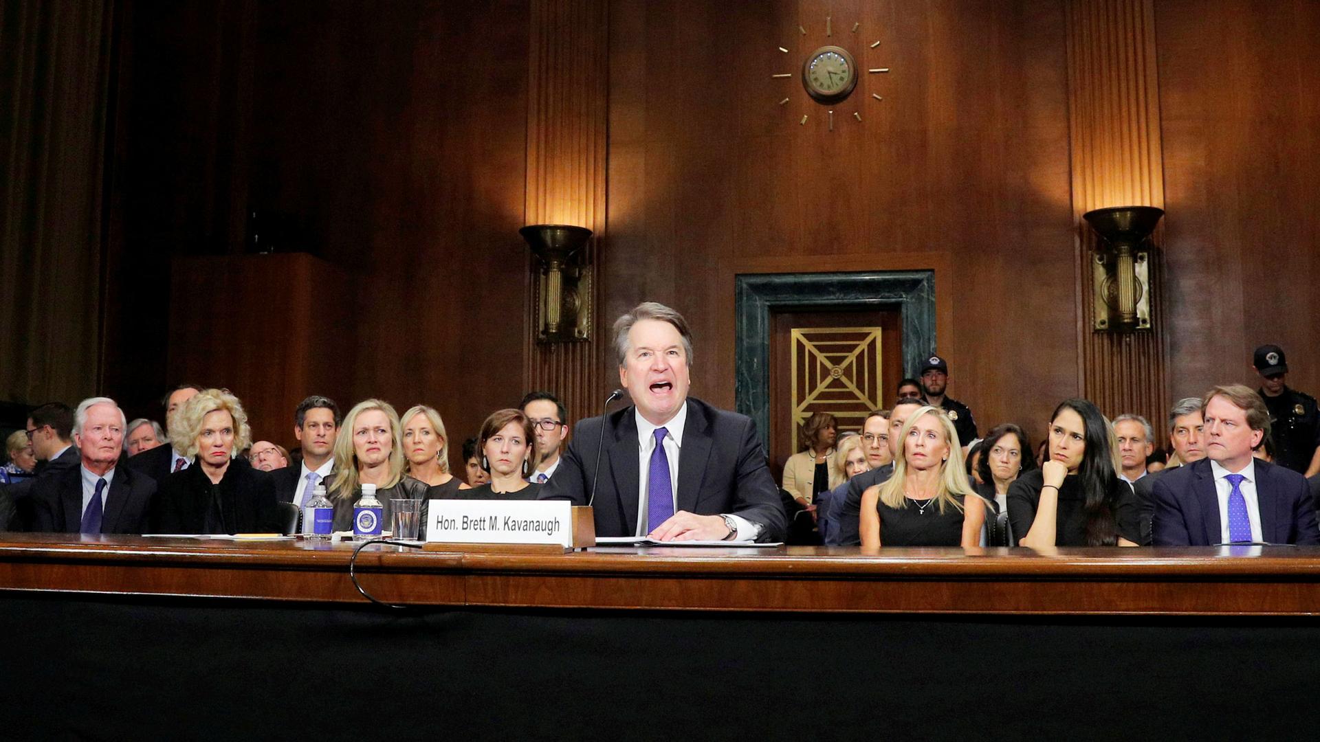 Brett Kavaugh is pictured at a table in the US Capitol. A row of women are behind him.