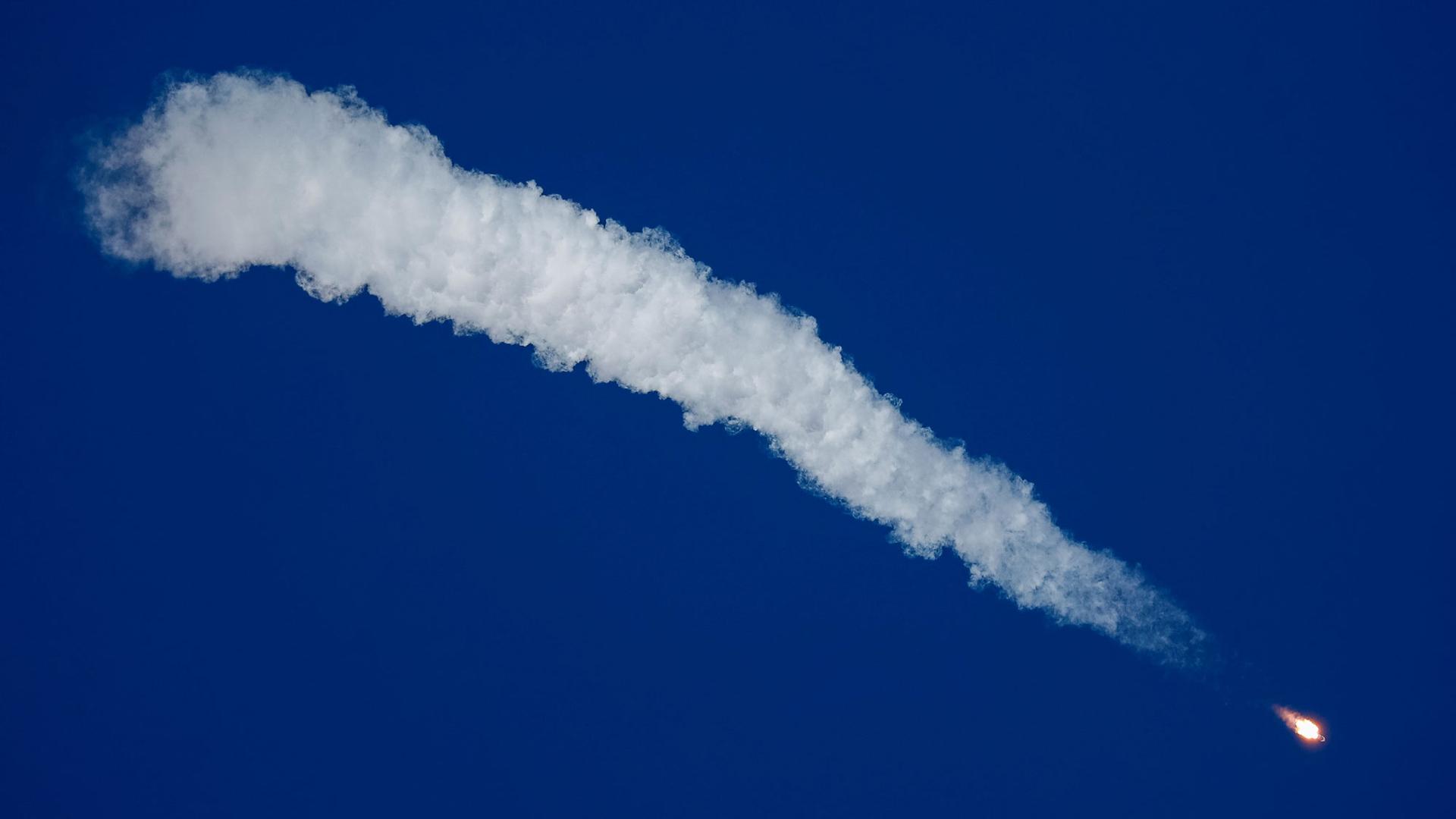 The Soyuz MS-10 spacecraft is shown streaking across the sky with a long white cloud of smoke trailing behind it.