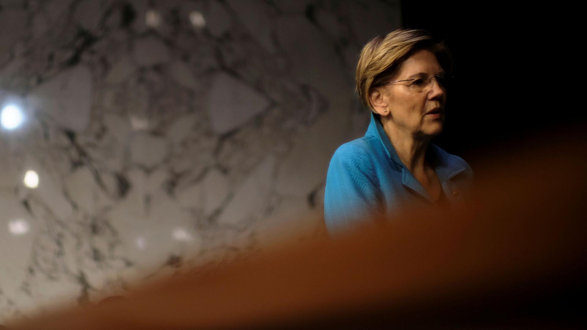 US Sen. Elizabeth Warren is shown wearing a blue jacket leaning against a marble wall.