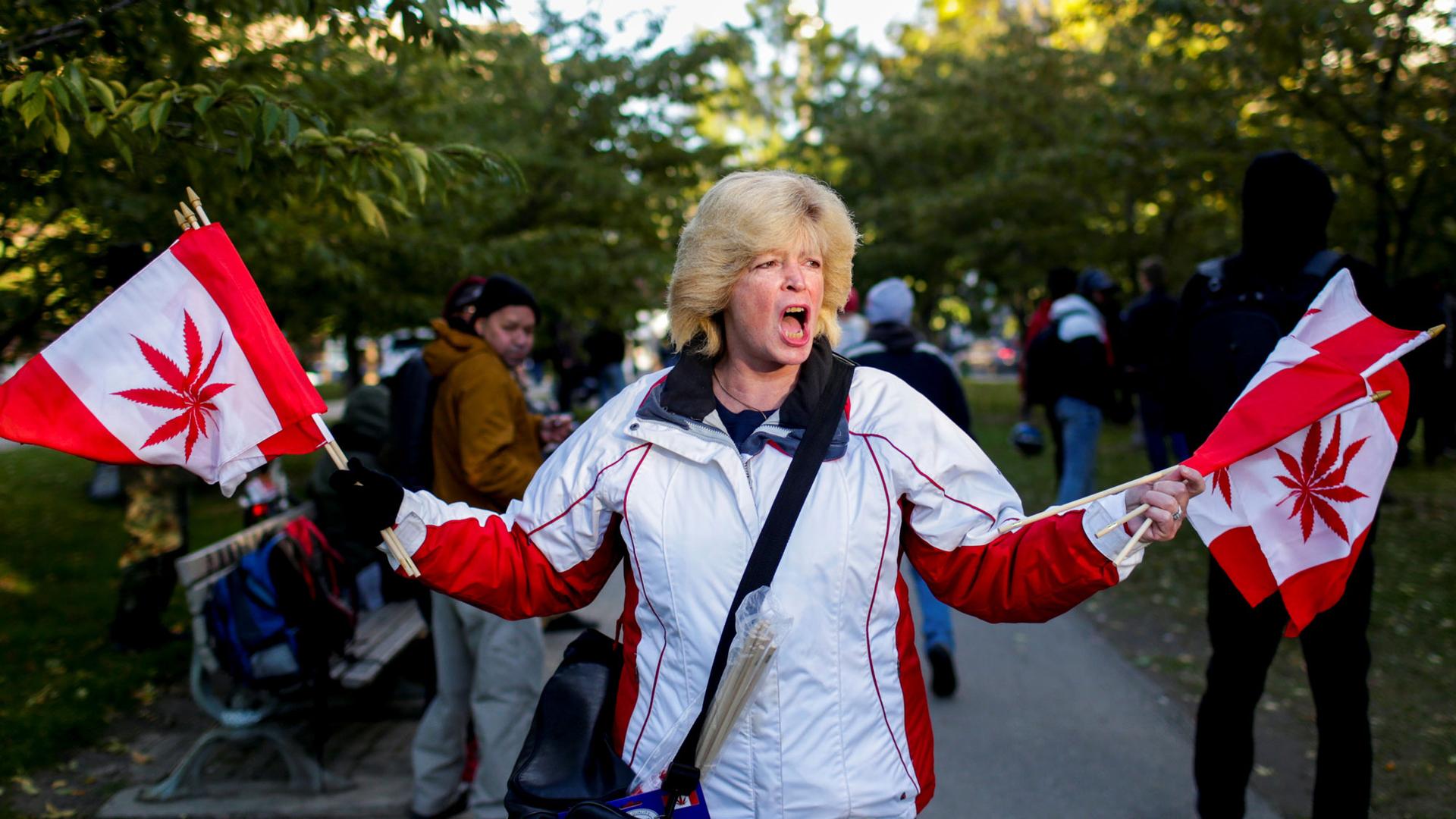 A woman is shown in a white jacket waving flags that resemble the Canadian flag but also include a maraijana leaf in the middle.