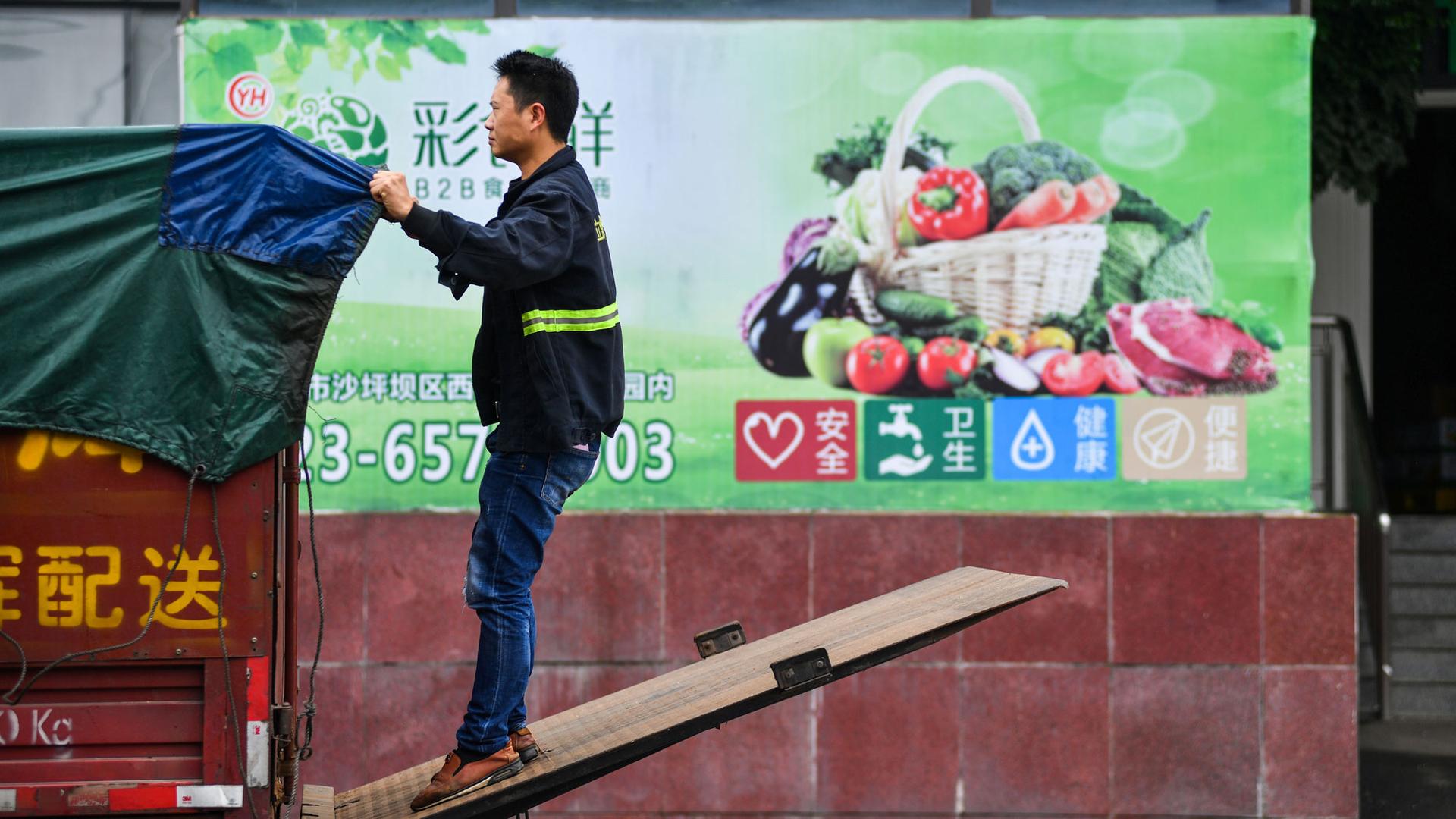 A worker wearing jeans and a reflective jacket adjusts a tarp on the back of a truck outside a warehouse of Yonghui Superstores in Chongqing, China.