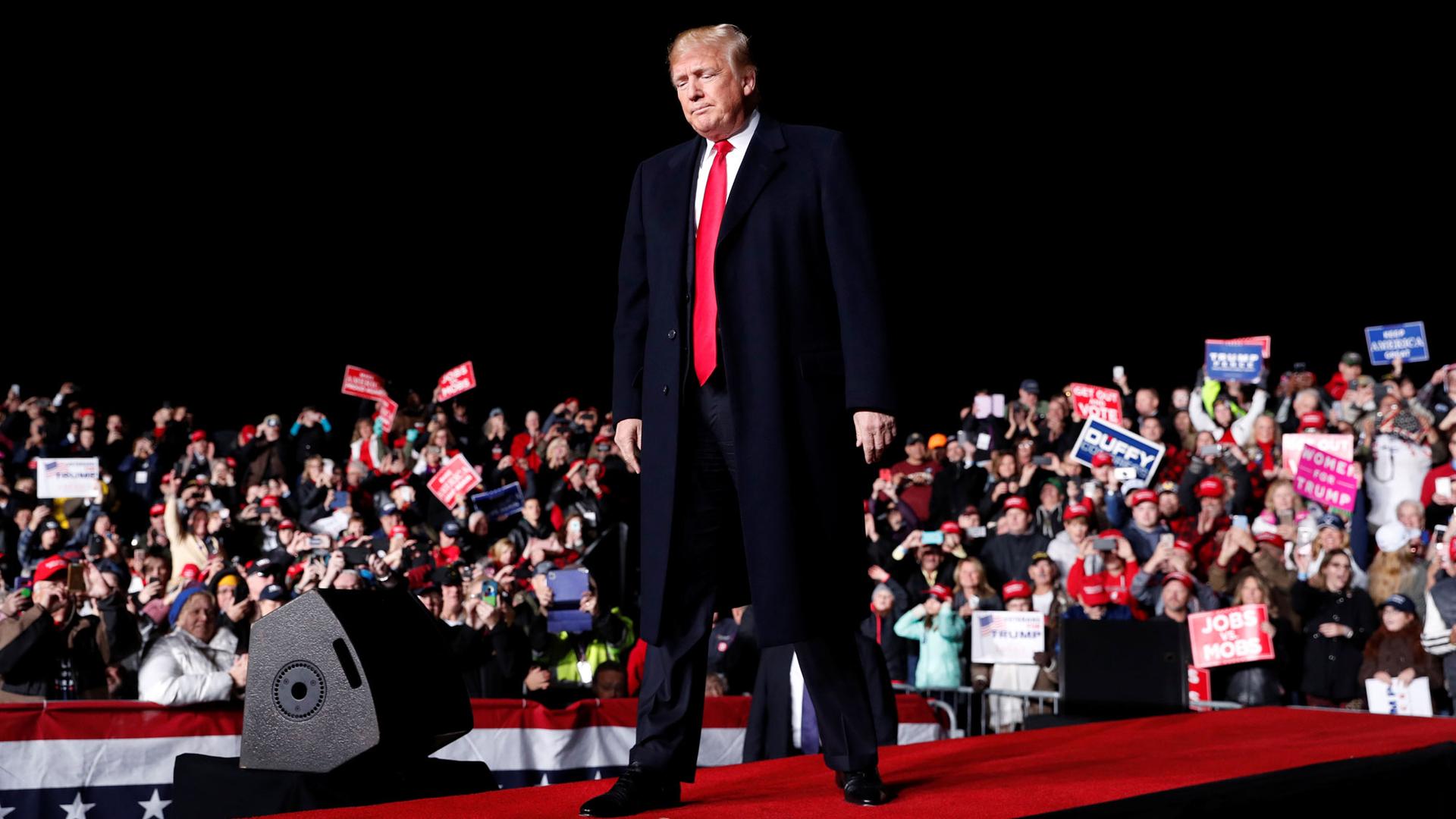 US President Donald Trump standing on a red carpet on a stage with a crowd behind him.