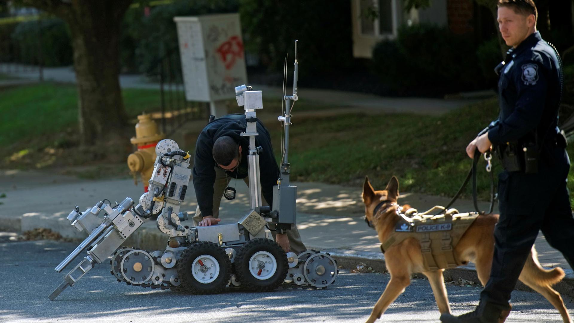 Law enforcement personnel walks with a dog while another bends over a bomb disposal robot.