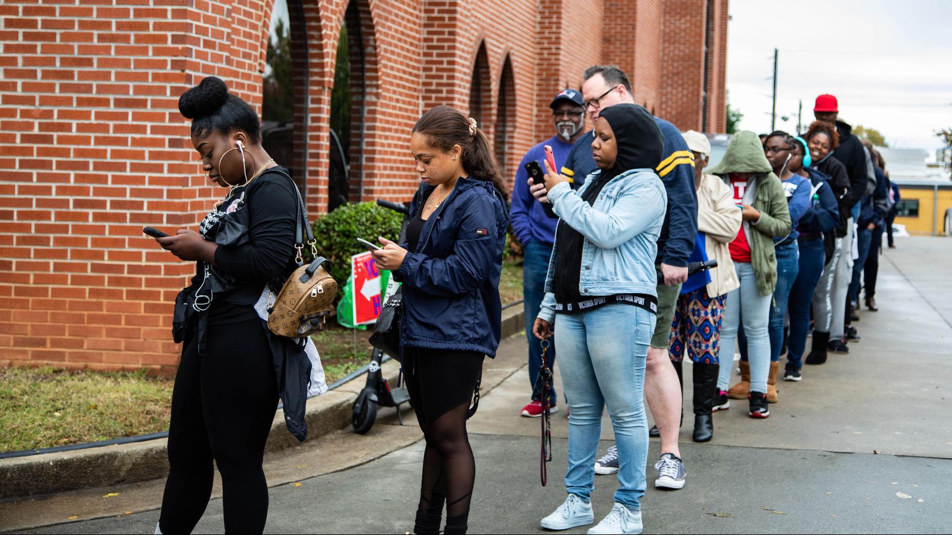 A line of people wraps around a brick building. Most people stand in line, looking at cell phones.