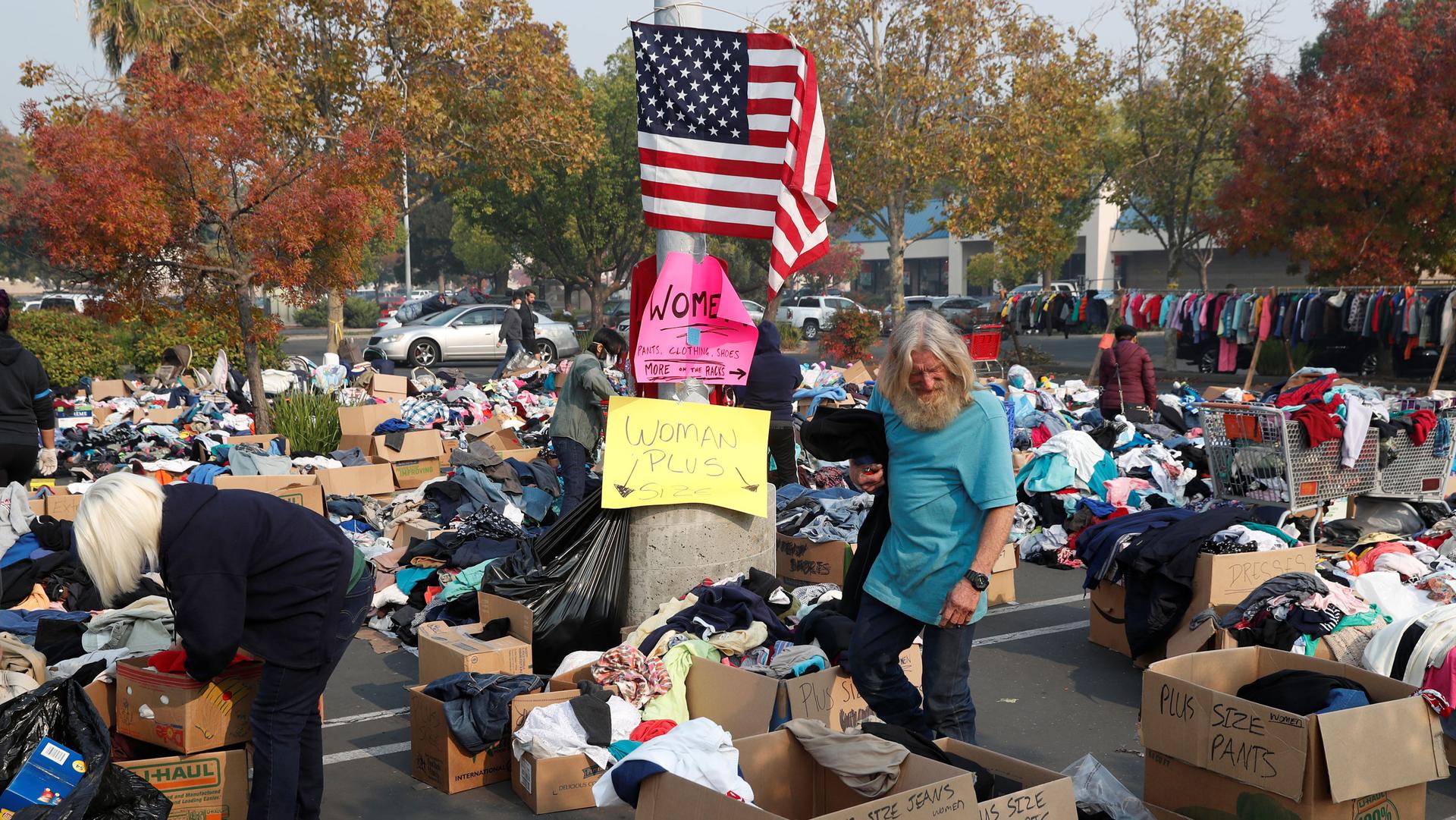 A man stands in a parking lot filled with boxes of clothes. On a lightpost, someone has taped up an American flag.