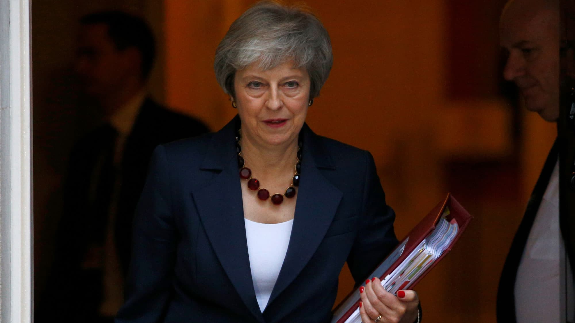 Theresa May is shown with a folder in her left hand walking out of 10 Downing Street, in London.