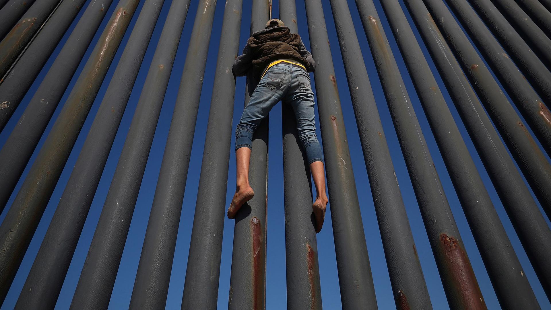 A person clings to a metal fence set against a bright blue sky.