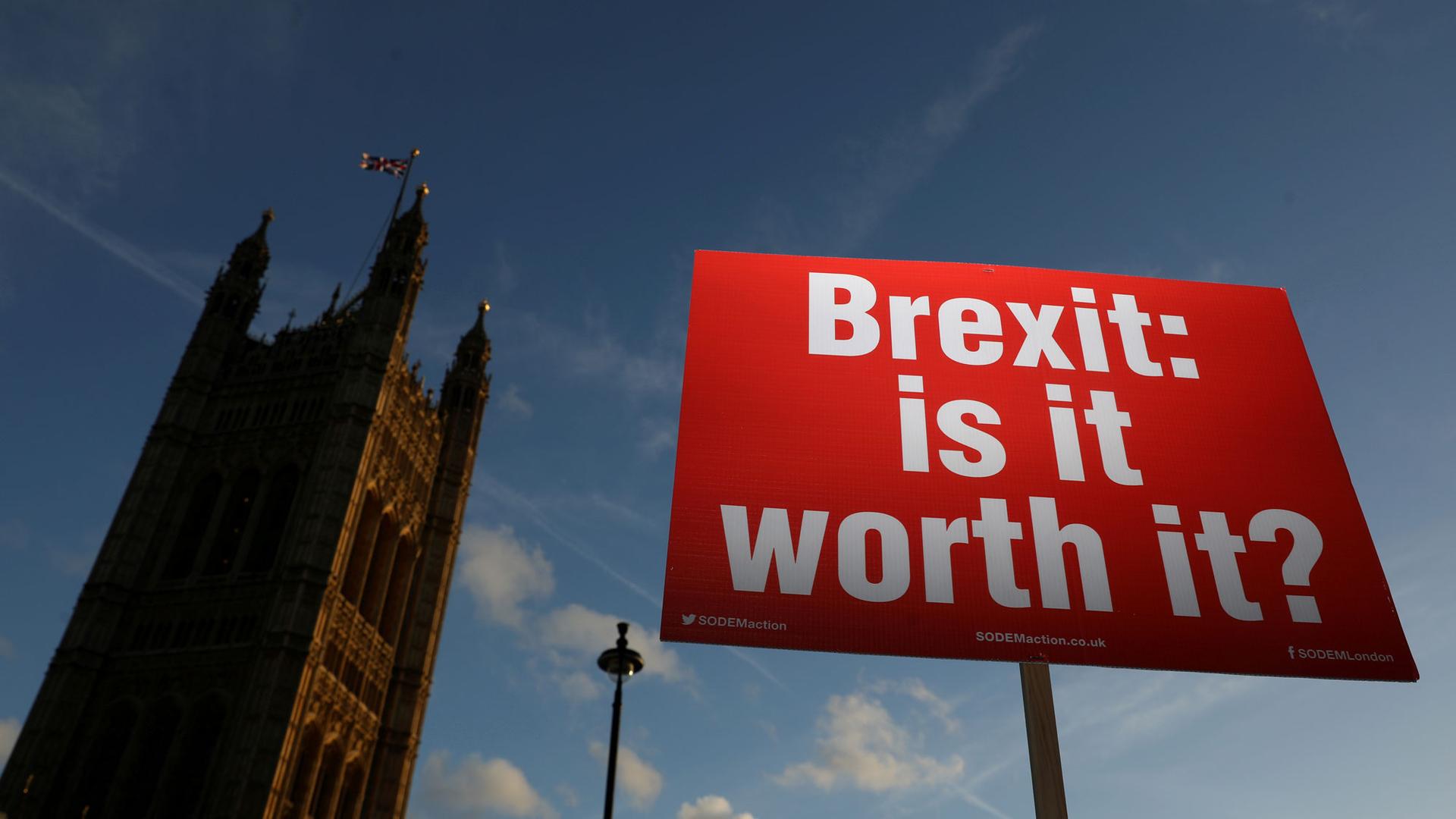 A red placard with white lettering reading, "Brexit: is it worth it?" is shown in the foreground with the Houses of Parliament in the background.