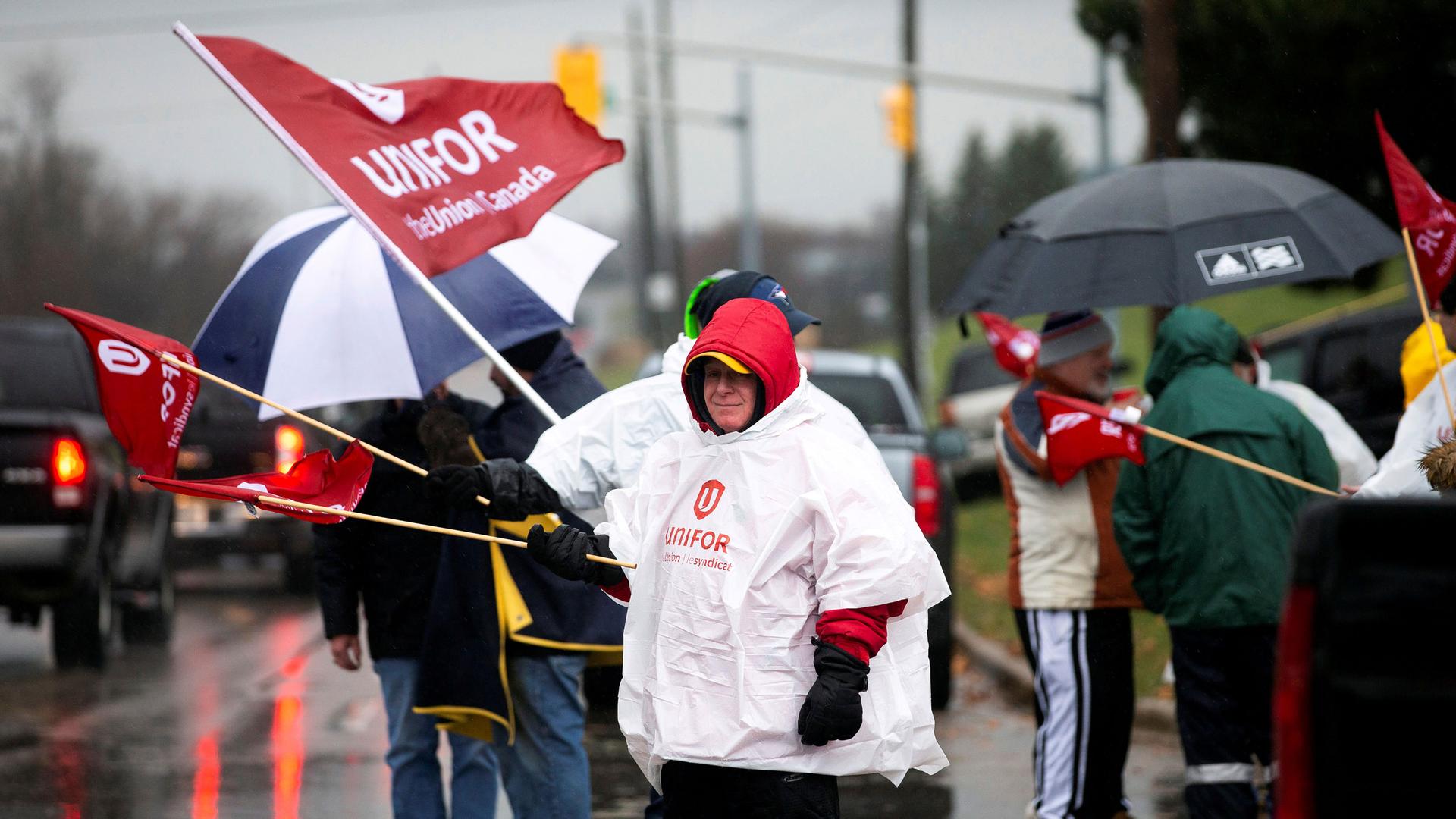 A person waves a red flag with the words "Unifor" on it with a group of others alongside a road.