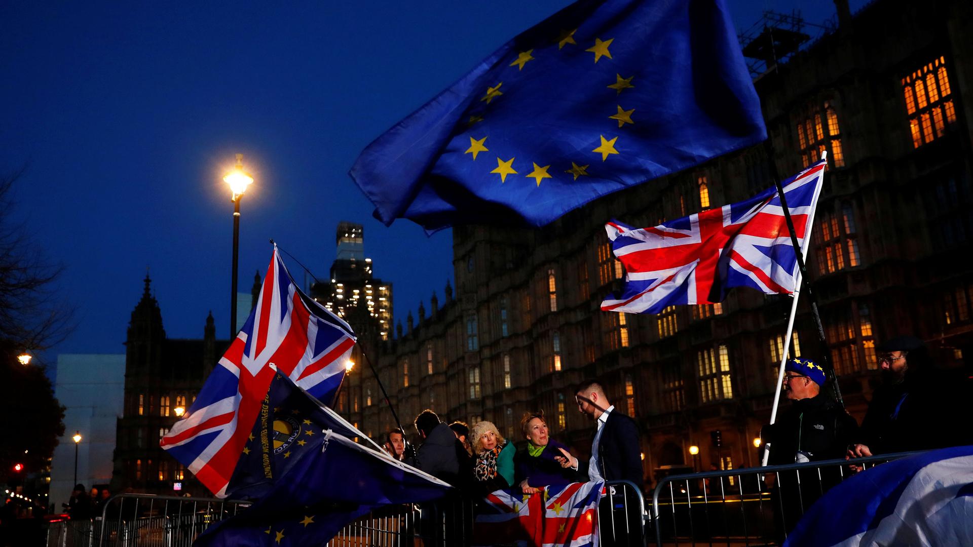 British flags wave against a dark sky outside the Parliament building in Long. People stand behind iron barricades holding the flags.