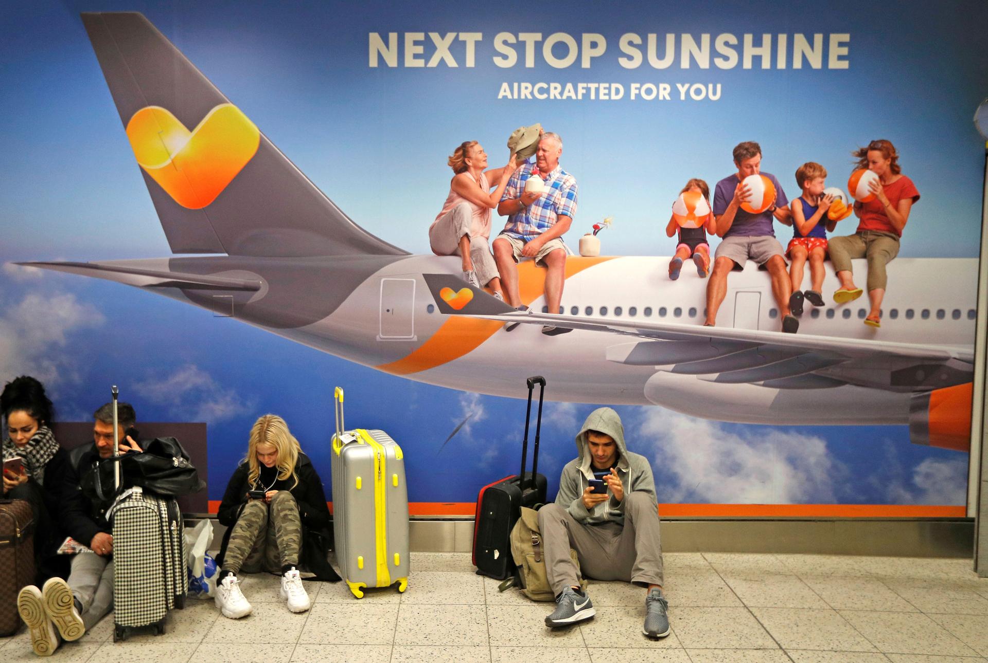 Airline passengers sit against a wall that is an advertisement for an airline that says 