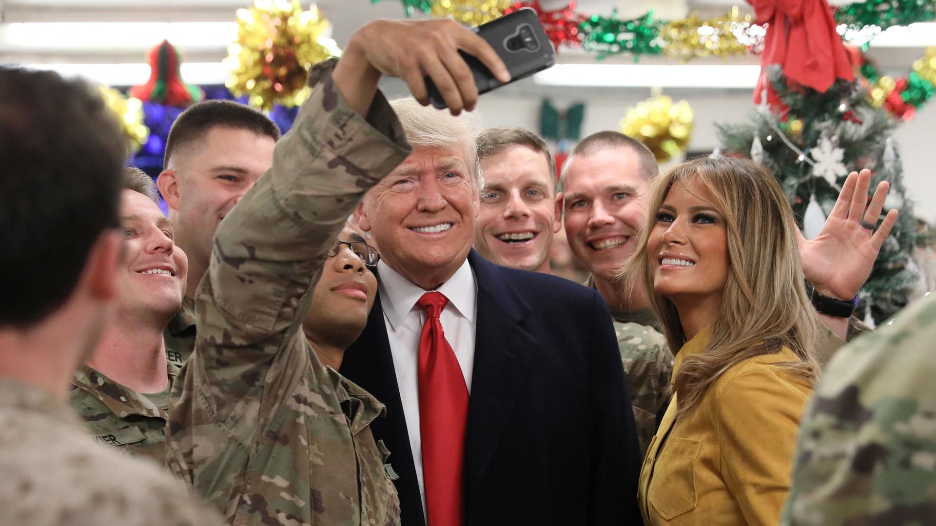 President Donald Trump and First Lady Melania Trump are shown surrounded by troops in camoflage while one soldier takes a selfie.