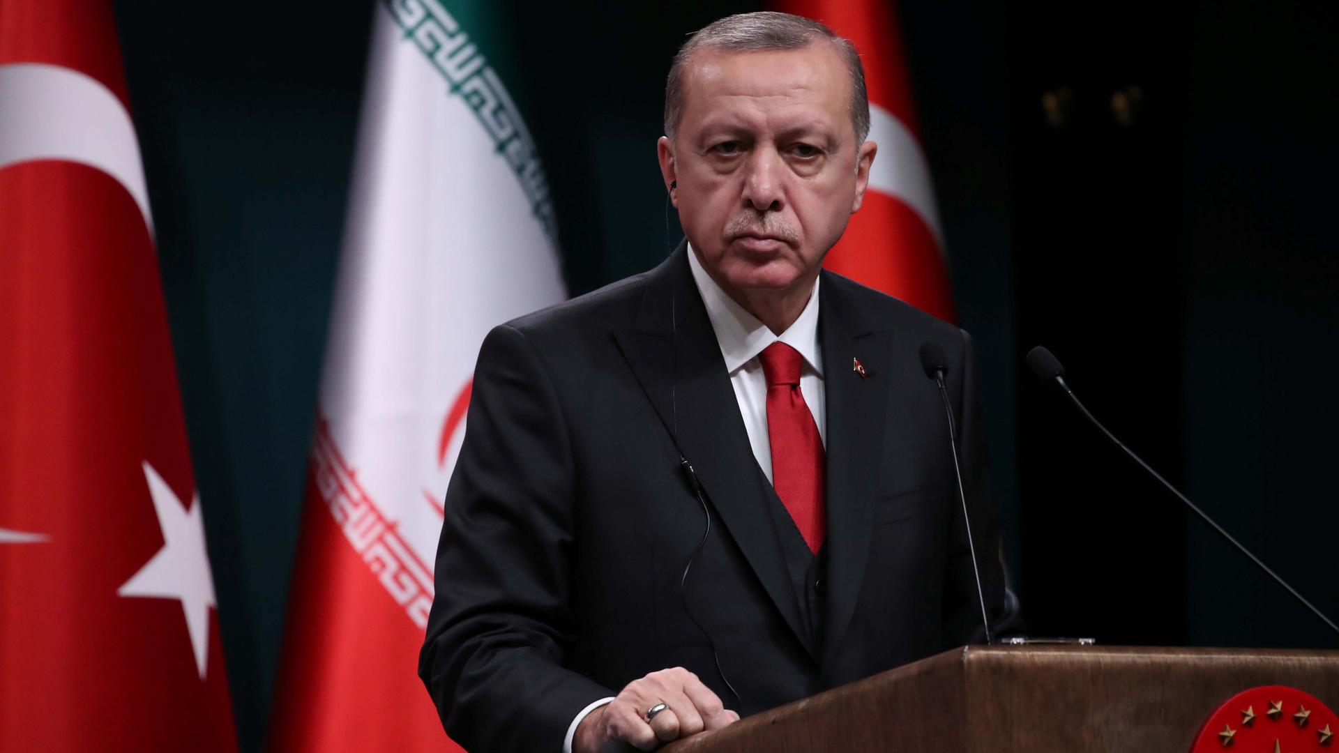 A middle-aged man stands at a podium with the Turkish and Iranian flag behind him.