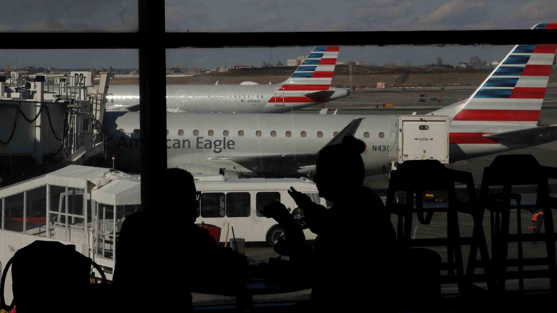 People waiting in an airport are silhouetted against big windows. Outside, planes are waiting next to gates.