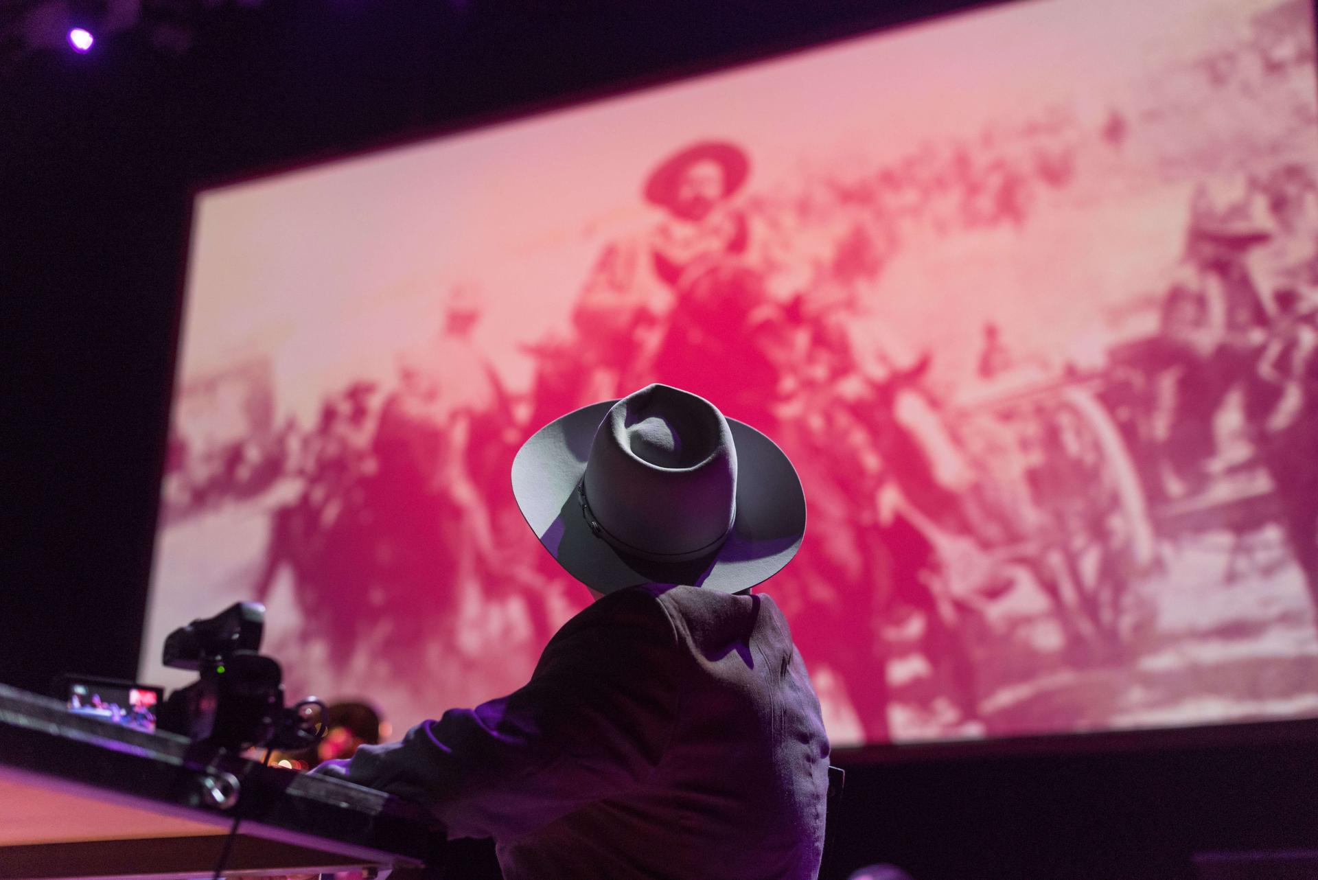 a performer wearing a hat looks at the giant projection of Pancho Villa on the stage film screen