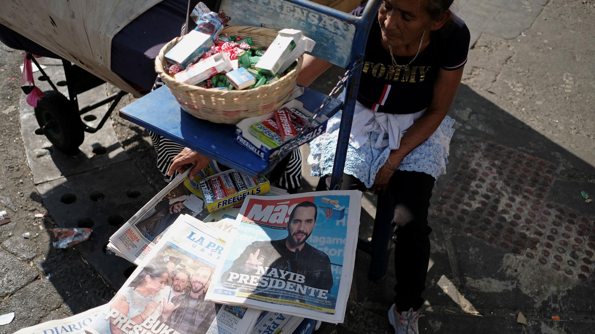 A woman sits on the street with piles of newspapers and gum for sale. The newspapers have the news that Nayib Bukele will be the next president of El Salvador.