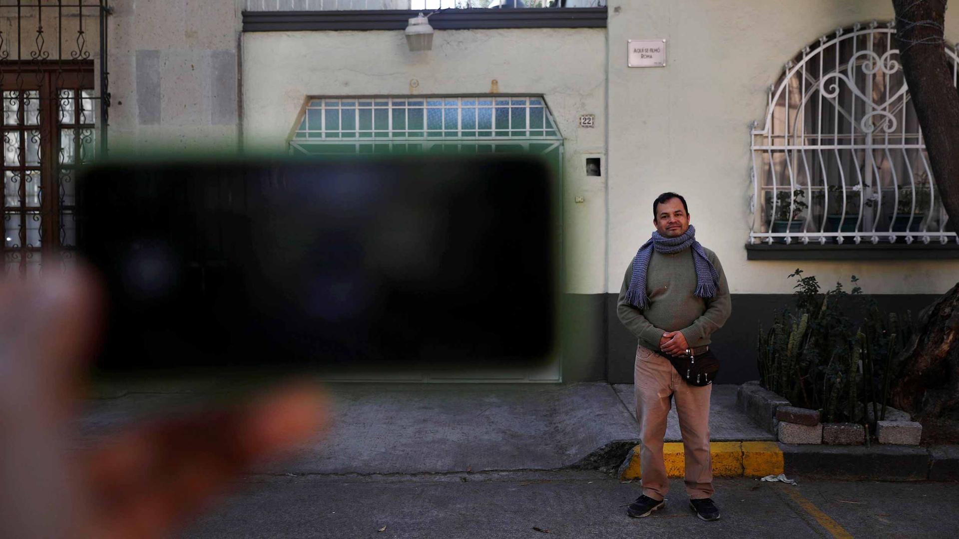 An iphone is blurry in the foreground as it takes a photo of a man standing outside a yellow home