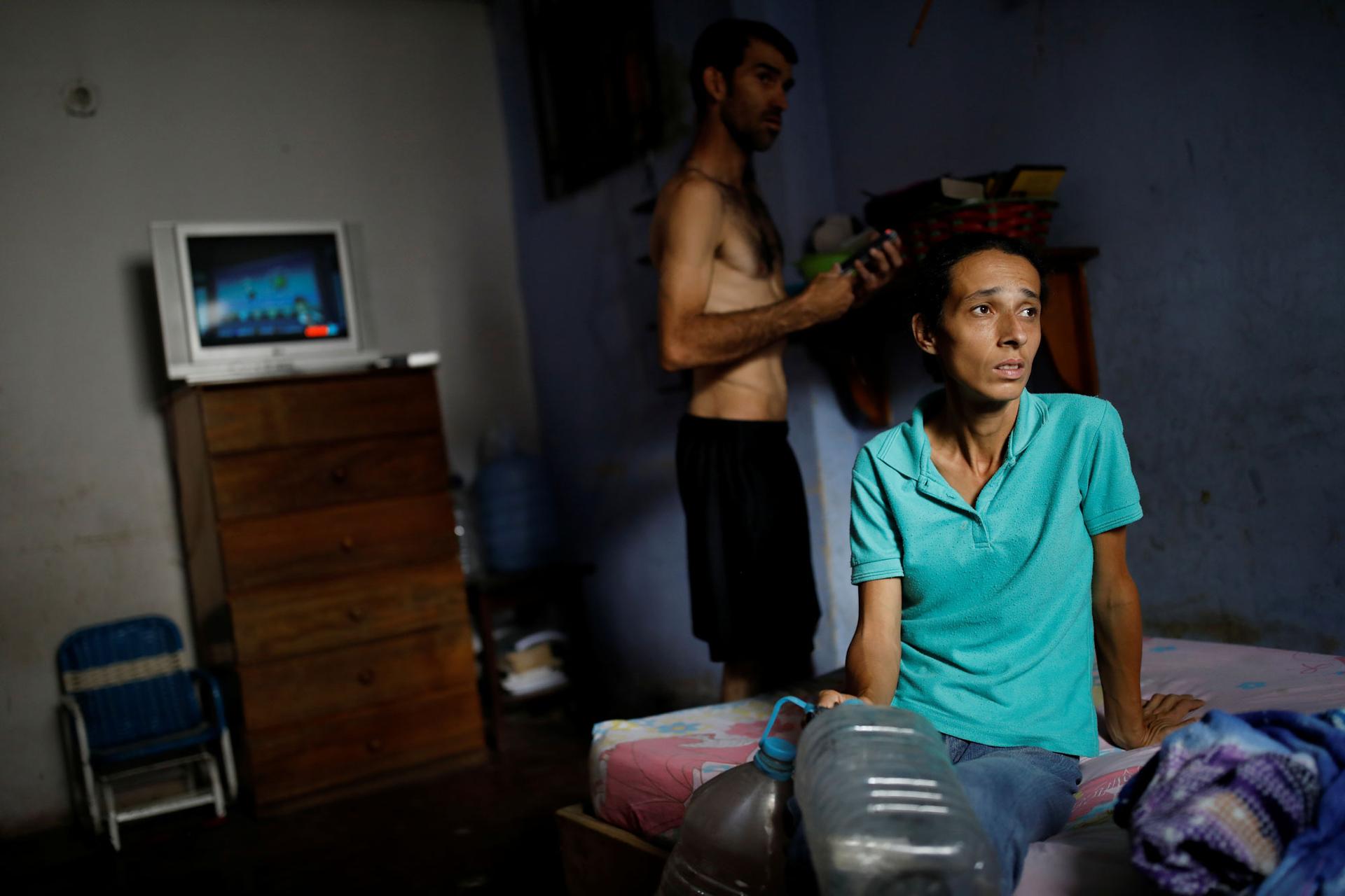 A woman is shown sitting a bed wearing a blue shirt as her husband stands behind her.