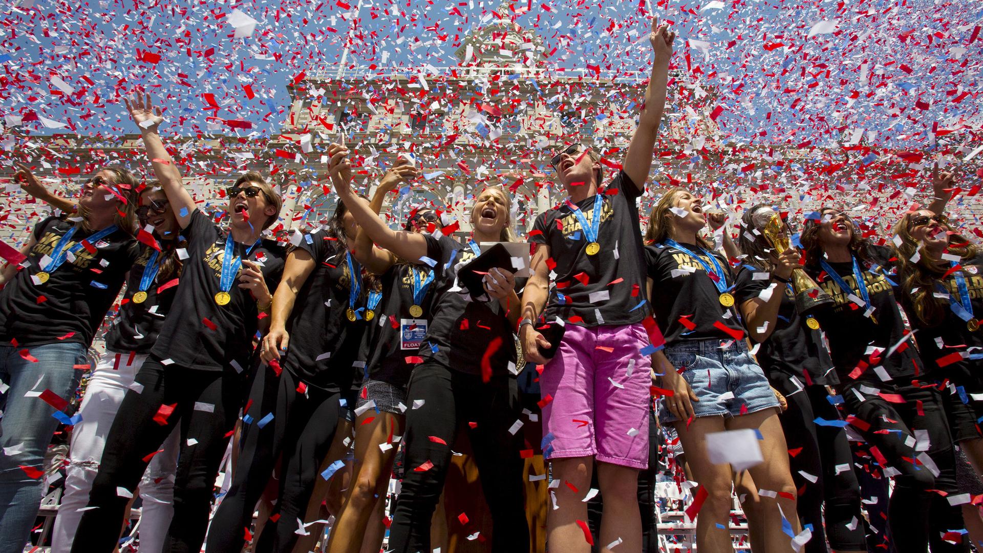 A crowd of women stand under a shower of confetti. They have gold medals around their necks and are wearing matching shirts reading "world champs."