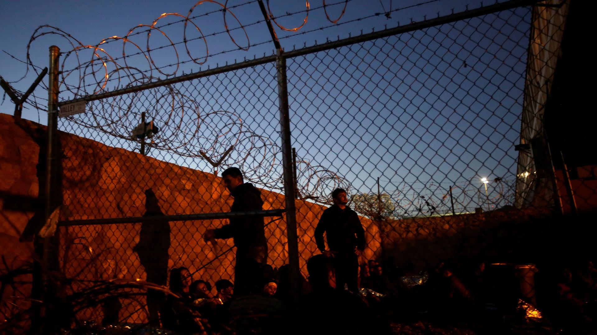 People a lit up by yellow streetlights as they sit inside a fenced area topped with razor wire as night falls