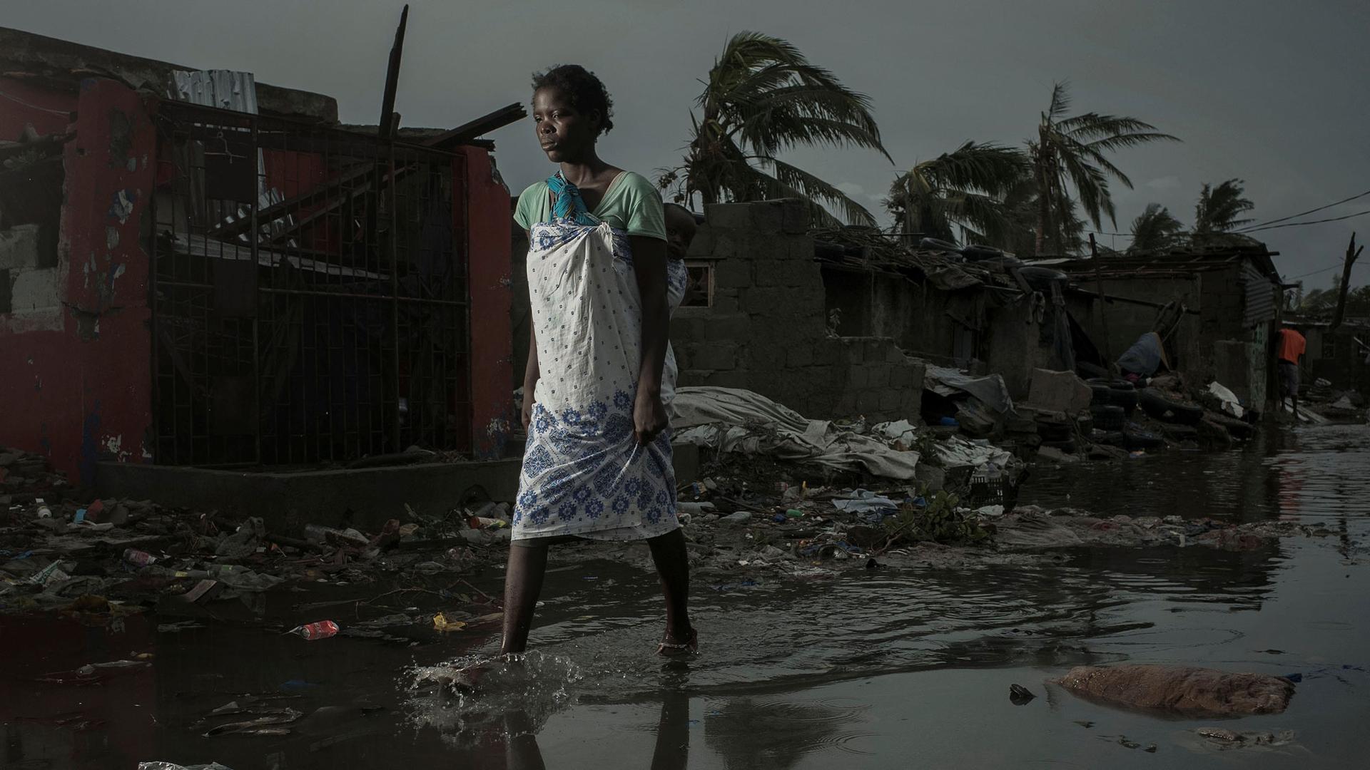 A woman is shown in ankle deep water walks down a flooded road with heavily damaged buildings in the background.