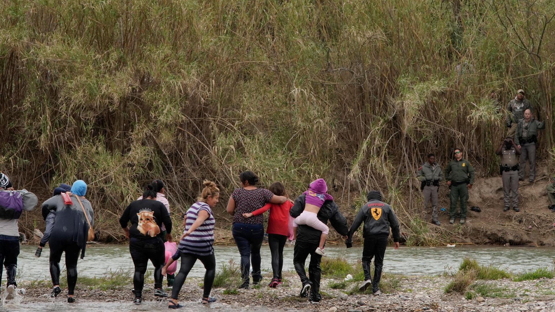 A group of migrants cross a shallow river and met with border agents in green uniforms.