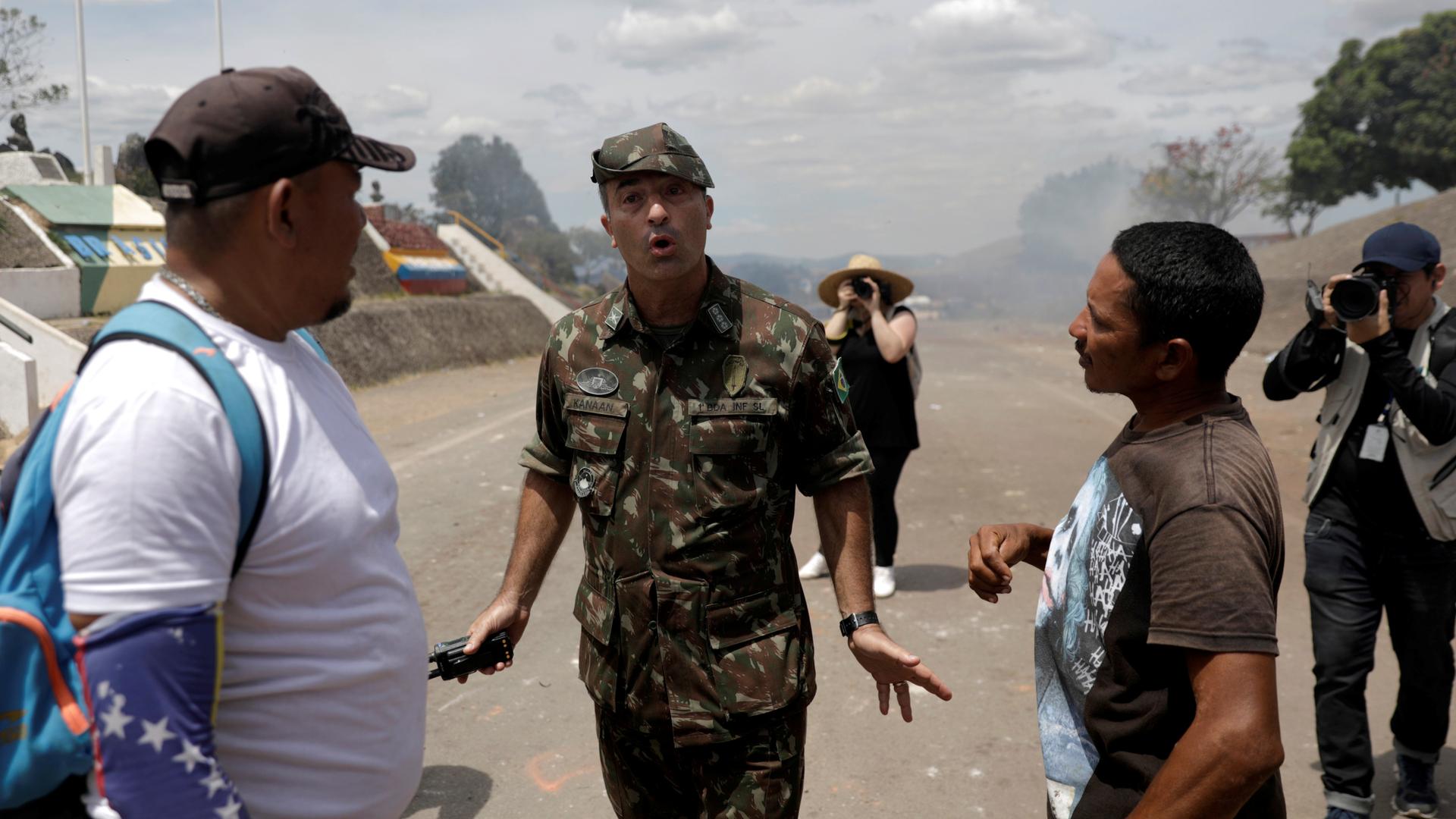 Man in uniform stands between two civilians with gesture of calm.