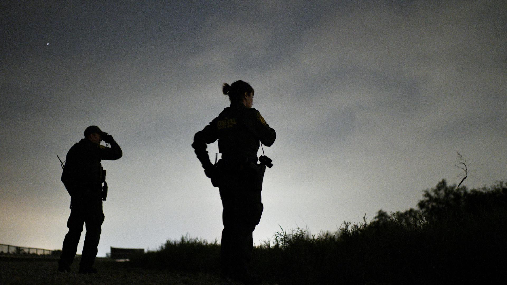 Two Border Patrol agents  are shown in silhouette looking off in a darkened field.