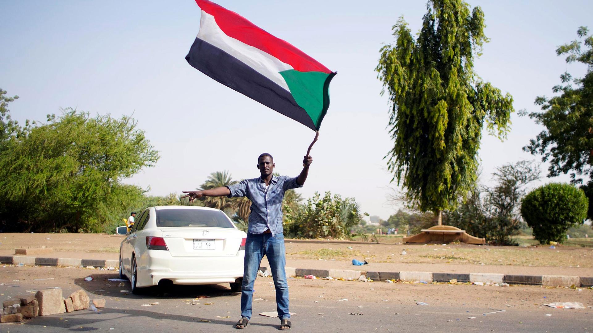 A man is shown wearing sandals, jeans and a button down shirt while waving the Sudanese flag.