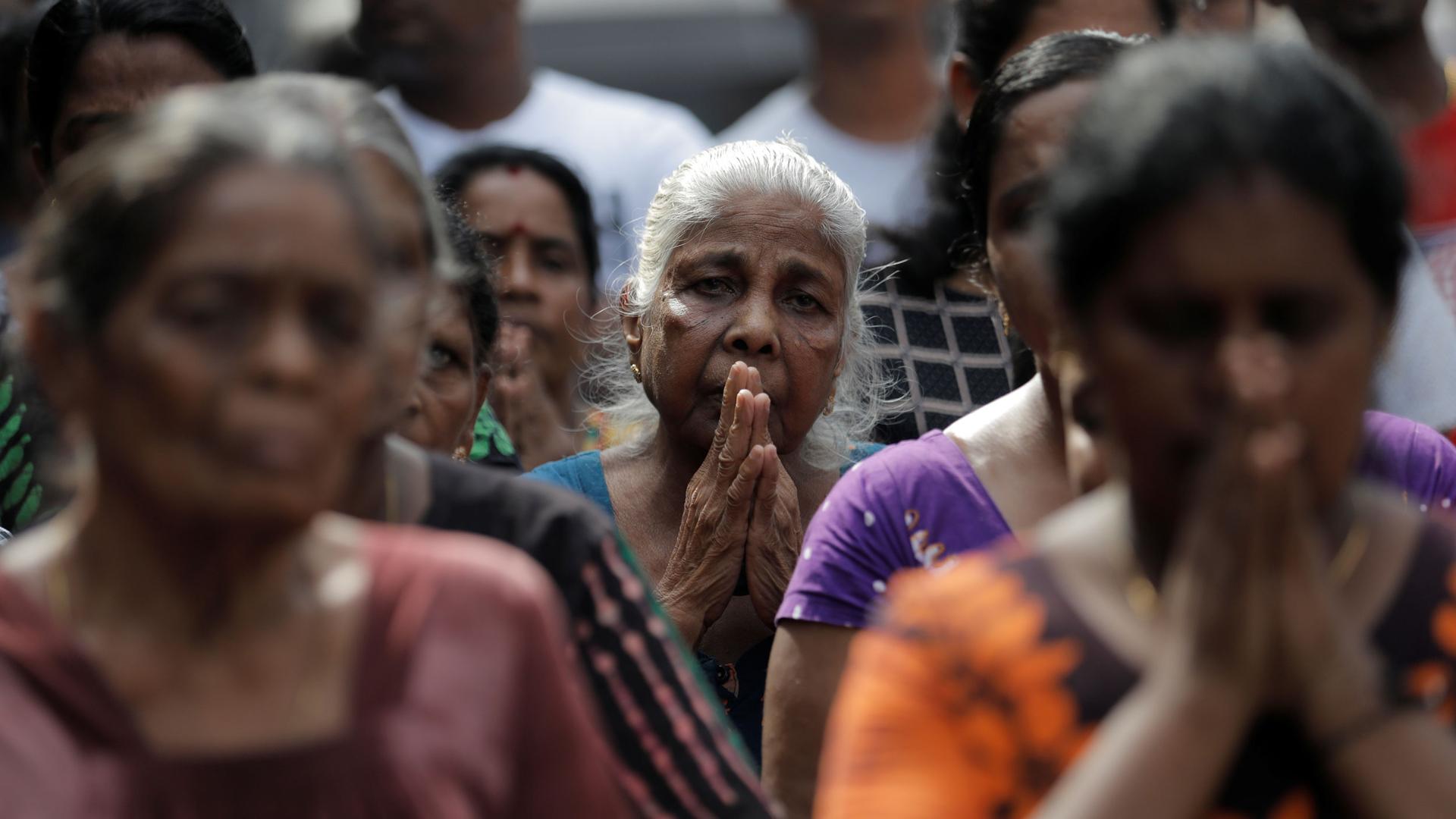 A crowd of older women are shown with their hands folded in prayer