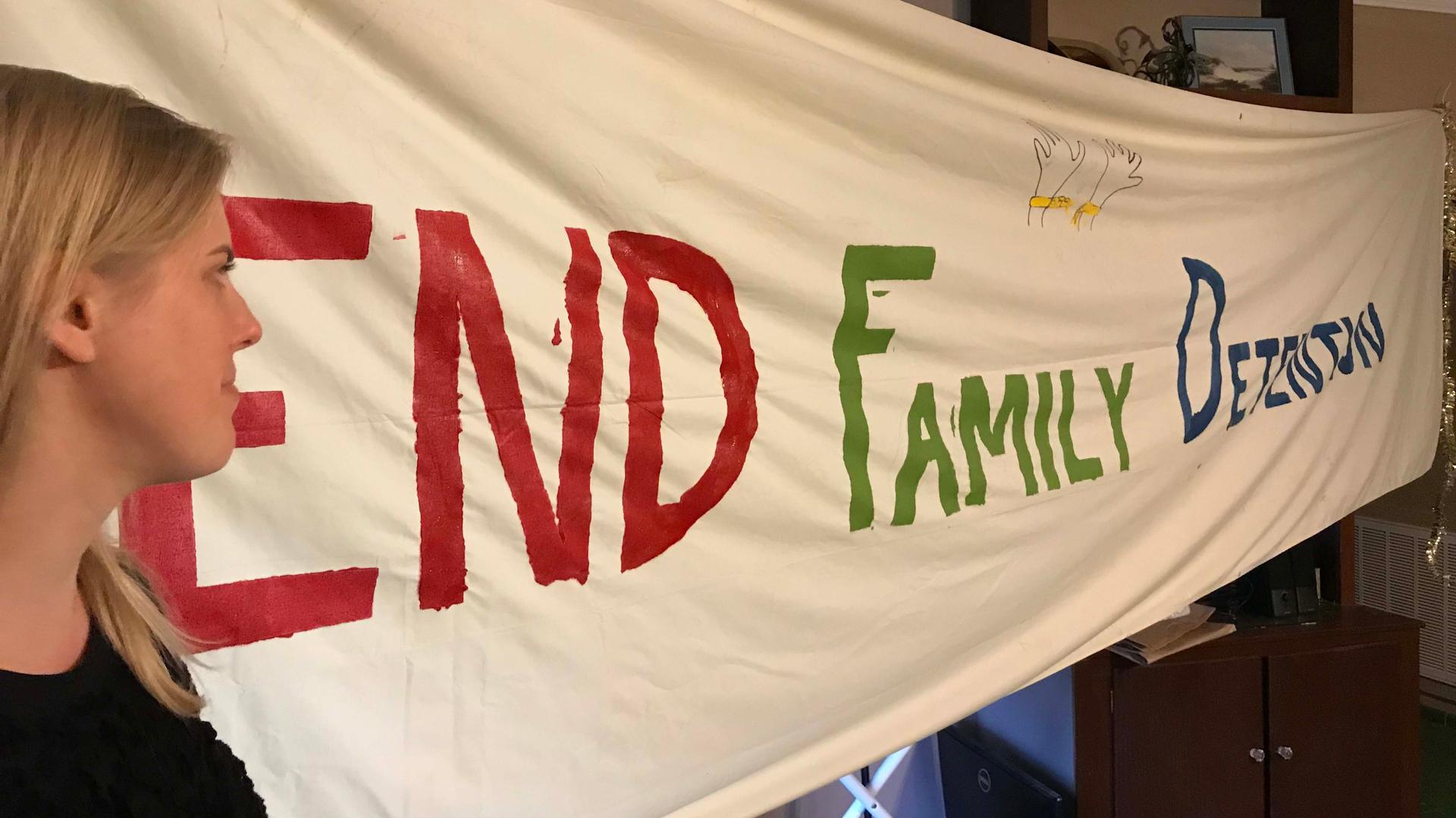 A woman stands in front of a banner reading "End Family Detention"